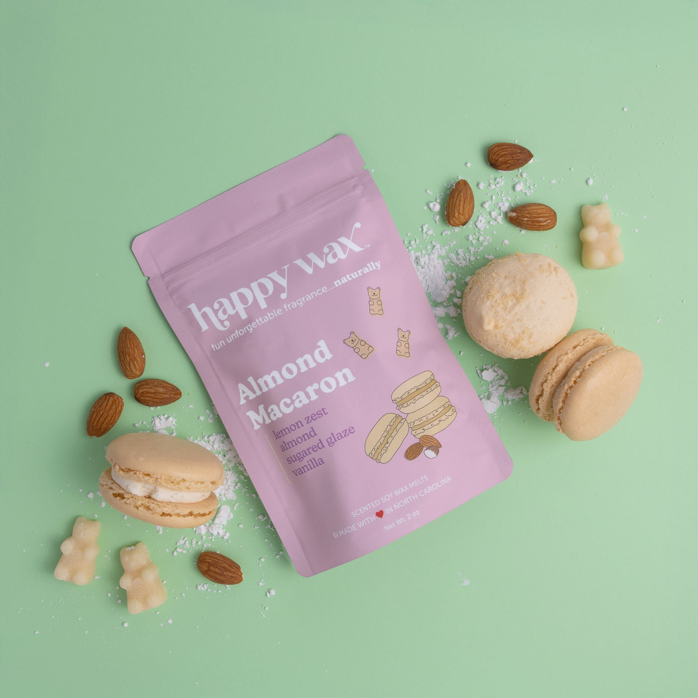 Pink pouch of Almond Macaron wax melts surrounded by almonds, macarons, and small wax bear shapes on mint background.