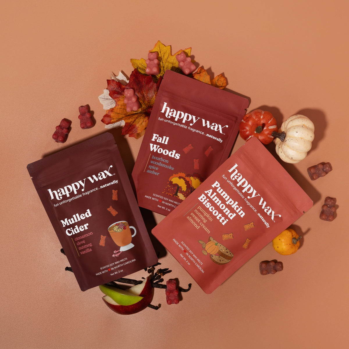 Three happy wax pouches in burgundy and coral with Fall Woods, Mulled Cider, and Pumpkin Birch scents surrounded by autumn le