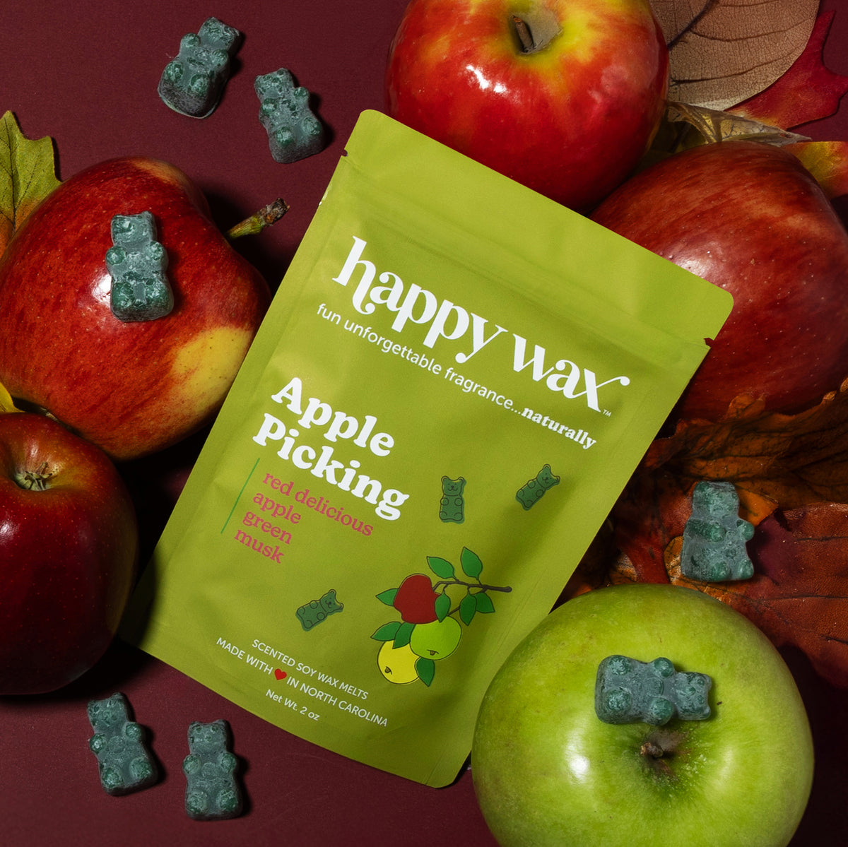 Lime green wax melt pouch surrounded by fresh red and green apples and teal bear-shaped wax melts on burgundy background.