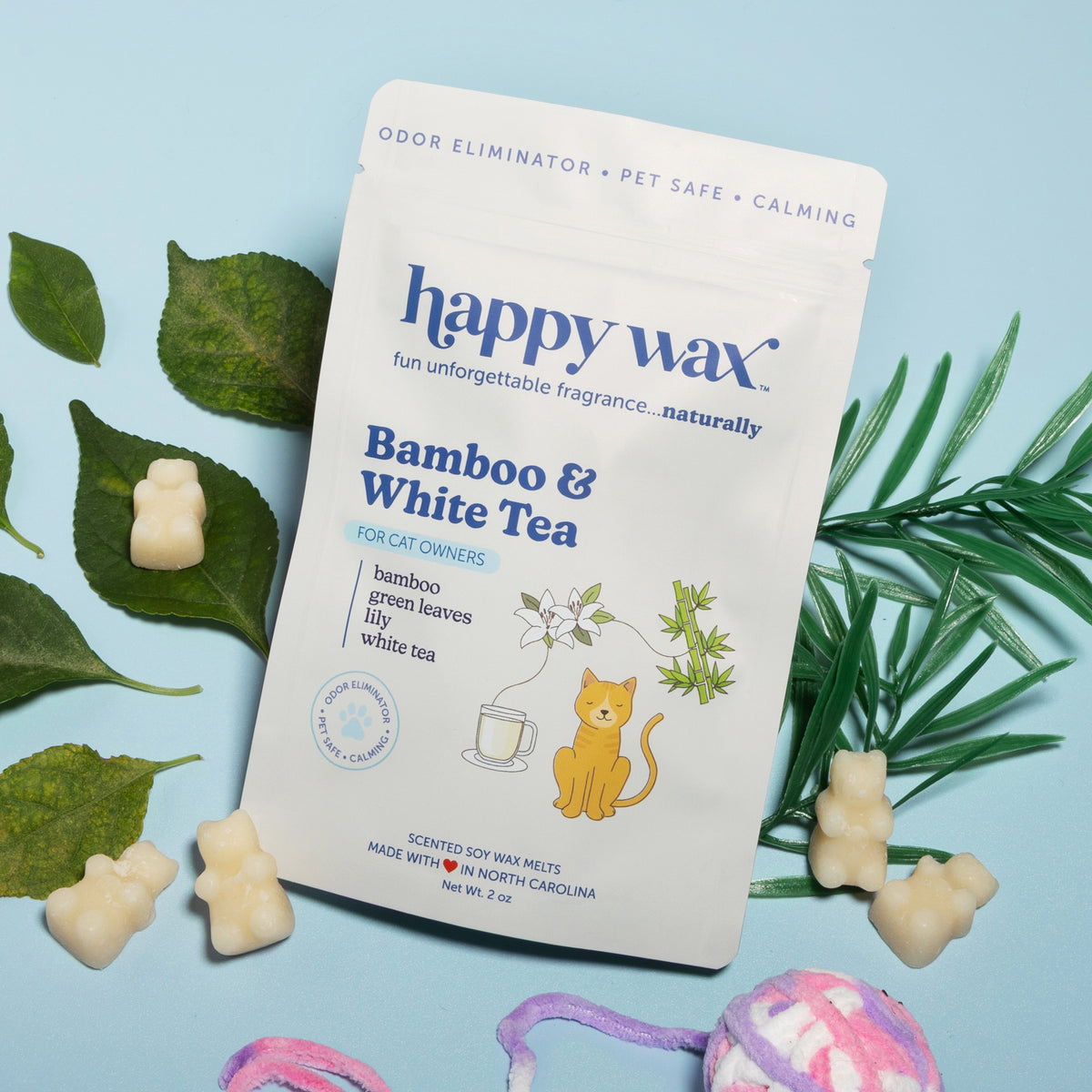 White package of bamboo and white tea scented wax melts surrounded by green leaves and cream-colored wax pieces on light blue