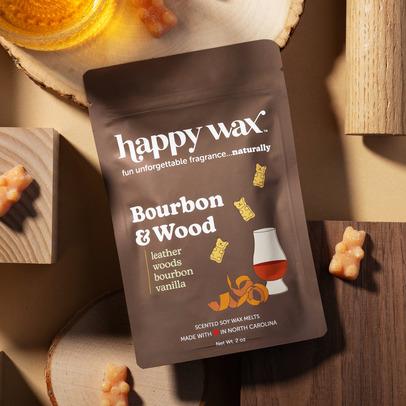 Dark brown wax melt pouch labeled "Bourbon & Wood" surrounded by wood slices, gummy bears, and a bourbon glass on wooden surf