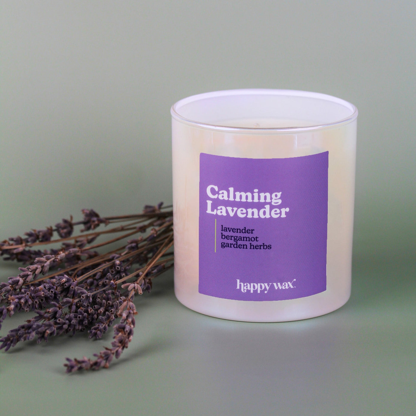 White cylindrical candle with purple label reading "Calming Lavender" next to dried lavender sprigs on neutral background.
