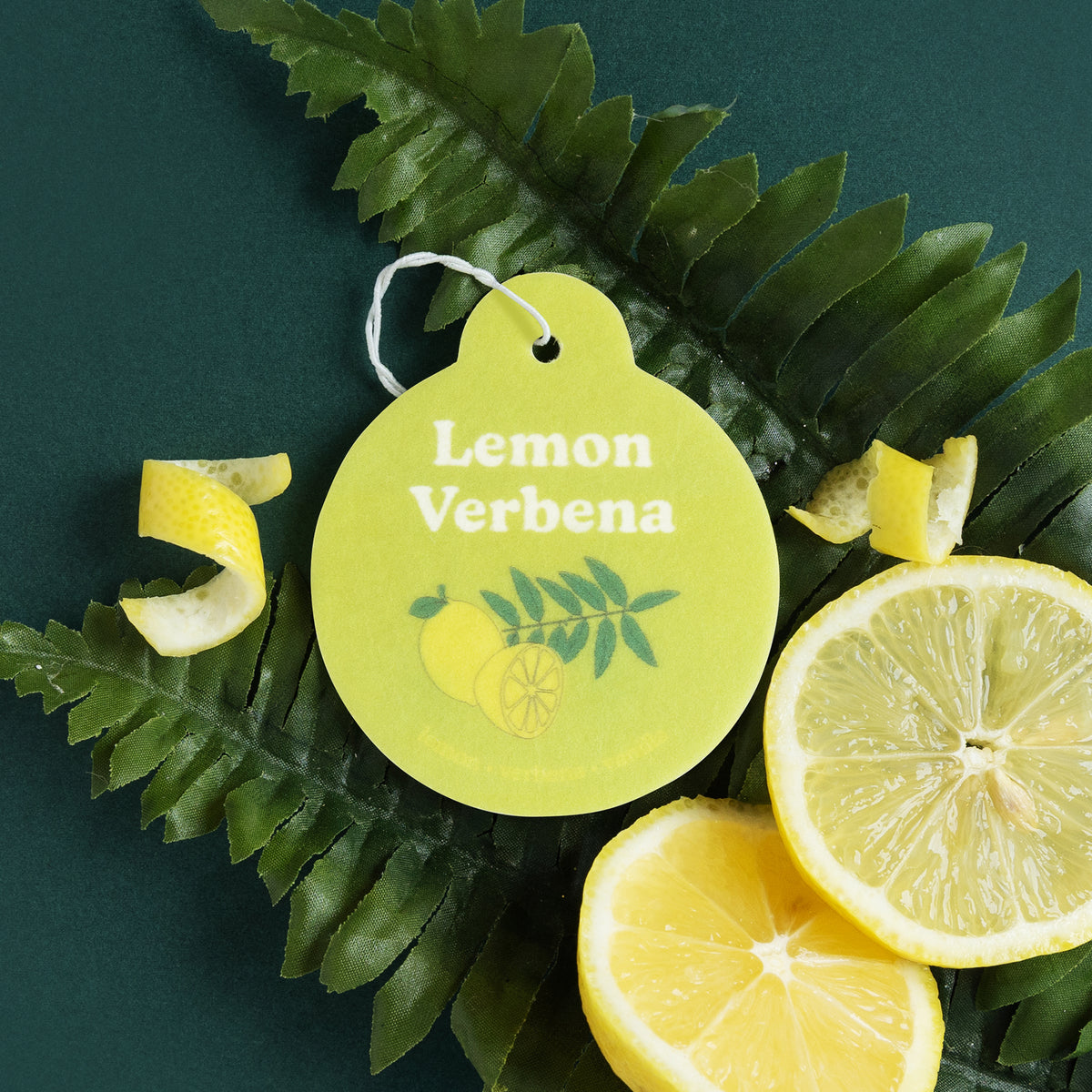 Yellow round car charm with "Lemon Verbena" text and leaf design, white string attachment, displayed with lemon slices and gr