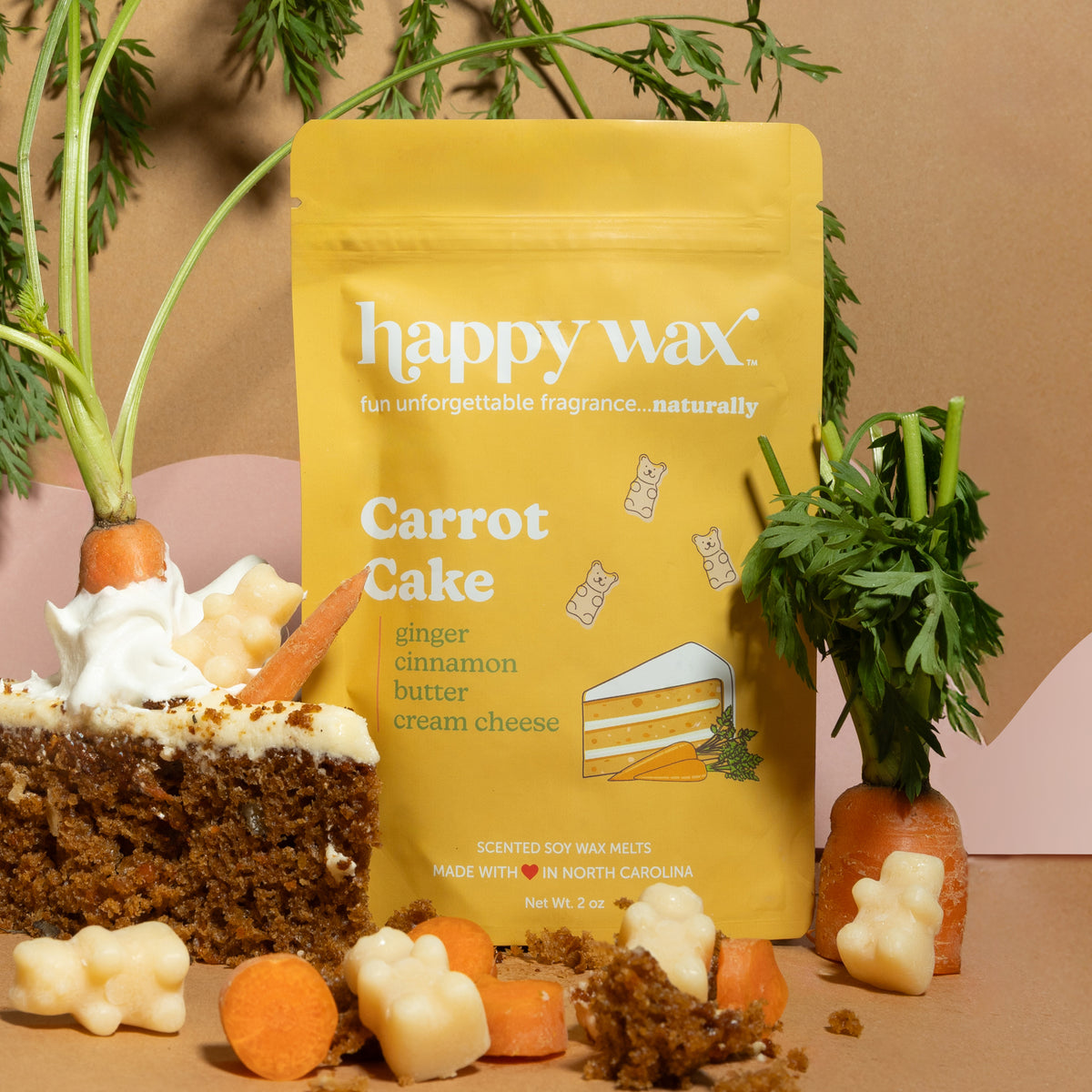 Yellow pouch of Carrot Cake scented wax melts surrounded by cake slices, carrot pieces, and fresh carrot greens on tan backgr