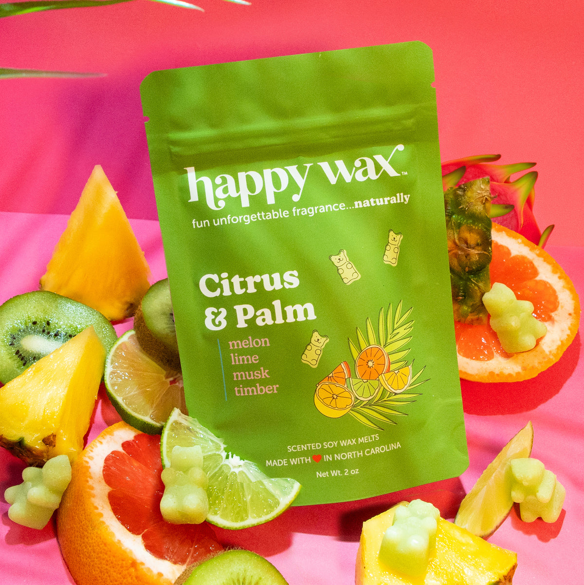 Green pouch of Happy Wax Citrus & Palm soy wax melts surrounded by citrus fruits and wax melts on pink background.