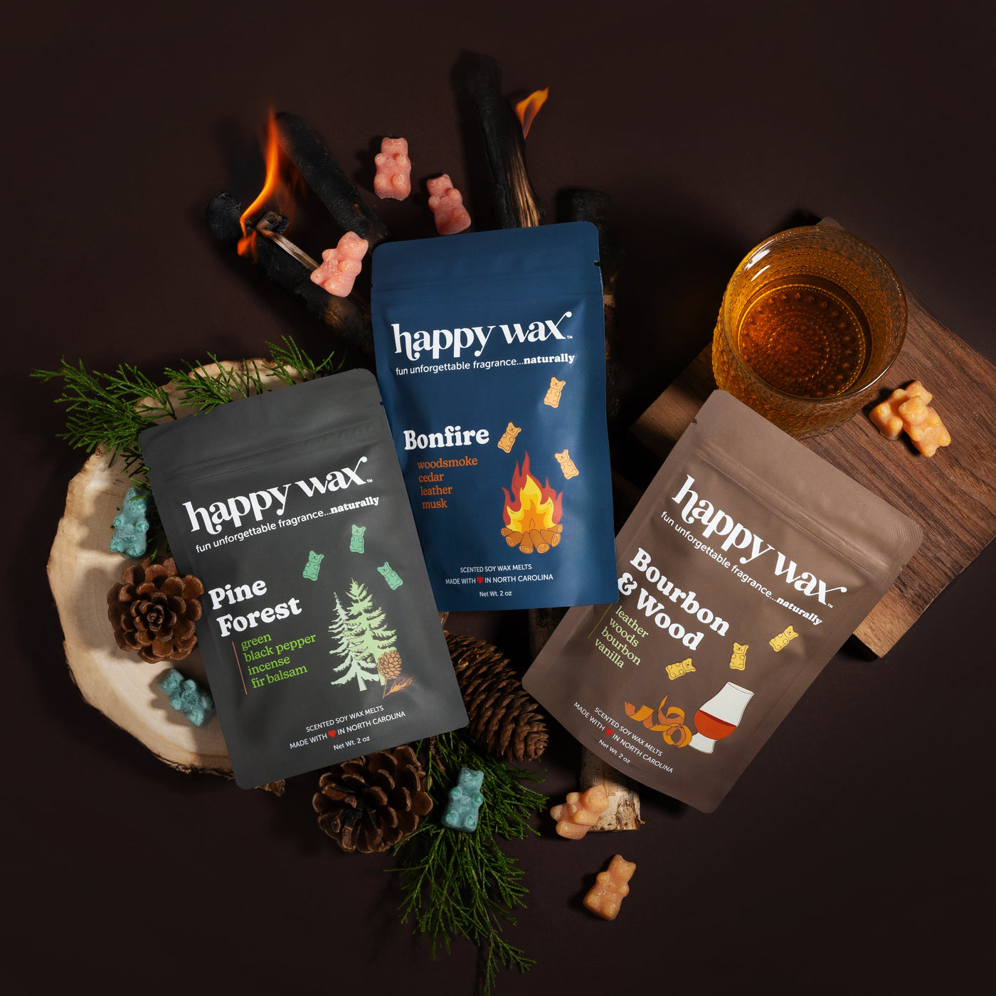 Three Happy Wax scented wax melt pouches in Pine Forest, Bonfire, and Bourbon wood scents displayed with pinecones, greenery,