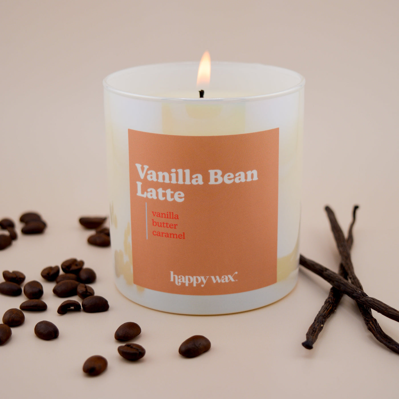 Cream-colored candle with orange label reading "Vanilla Bean Latte," surrounded by coffee beans and vanilla pods.