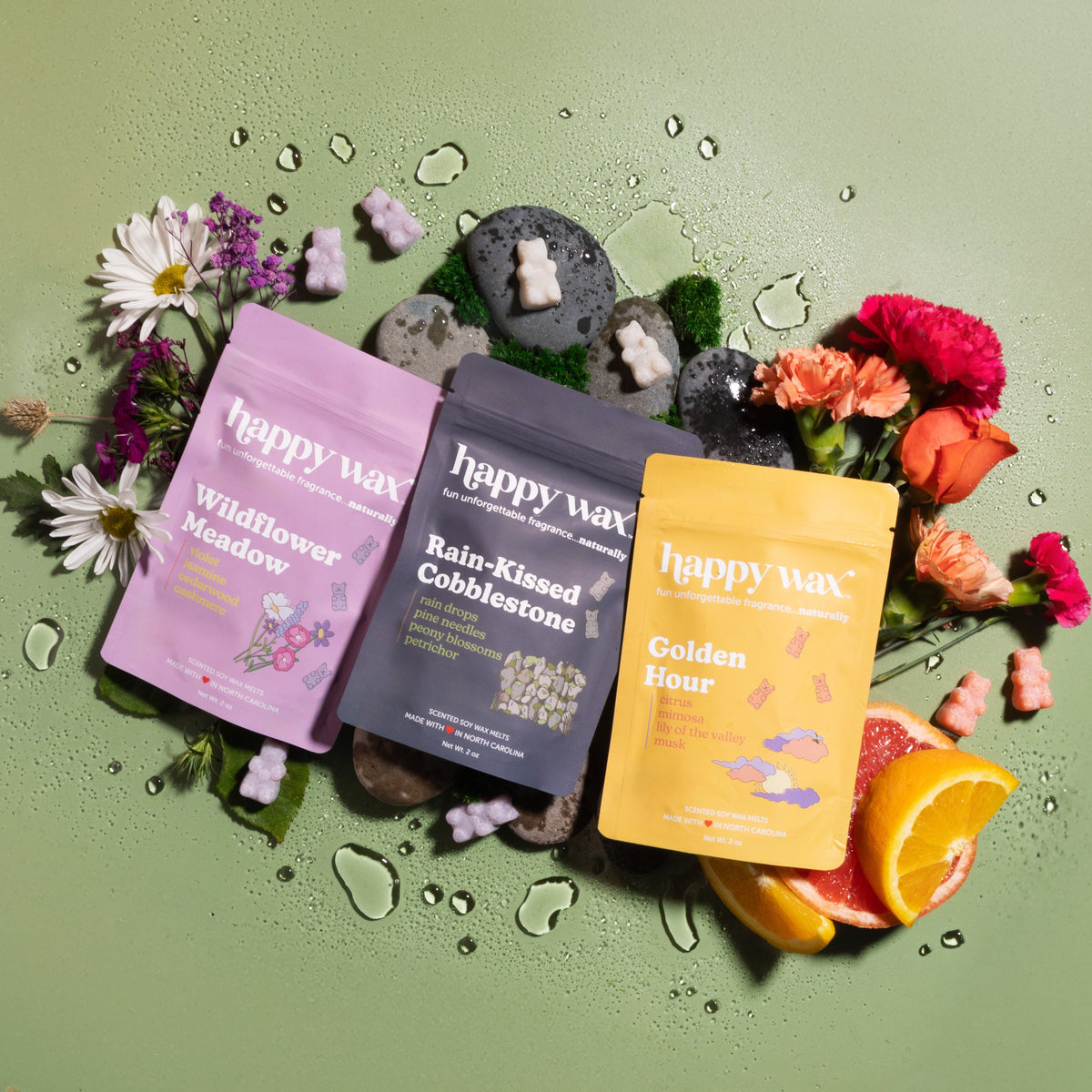 Three Happy Wax candle pouches in purple, dark gray, and yellow arranged with fresh flowers, stones, water droplets, and citr