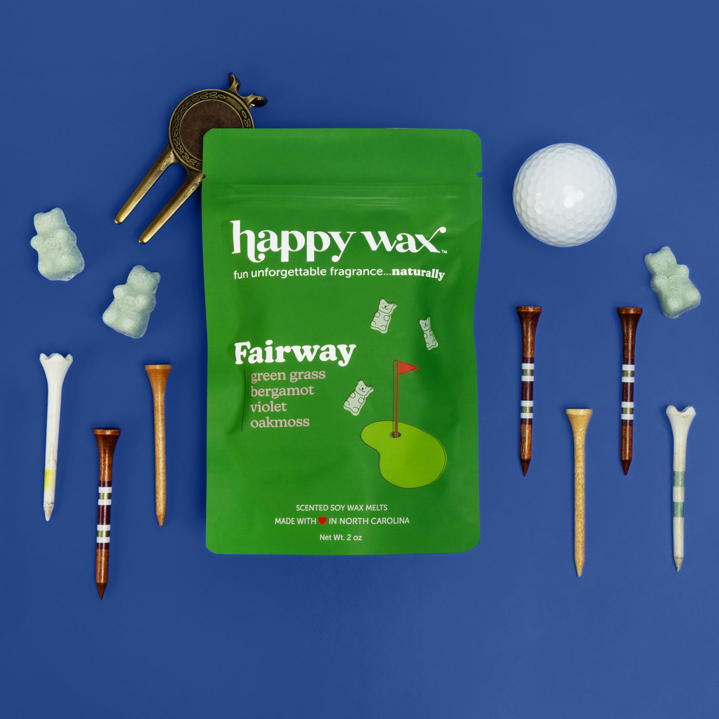 Green Fairway wax melts pouch surrounded by golf-themed items including a white ball, tees, and a brass divot tool on blue ba