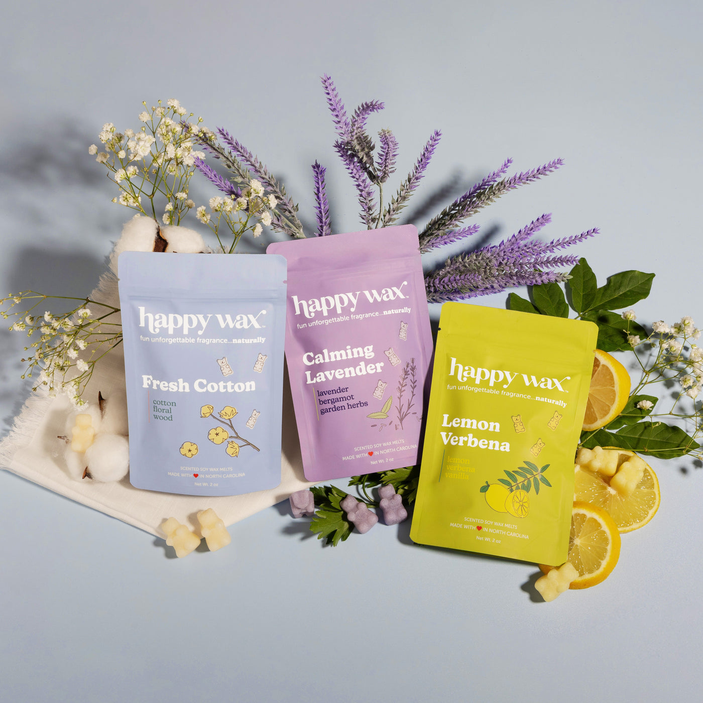 Three Happy Wax candles in blue Fresh Cotton, pink Calming Lavender, and yellow Lemon Verbena surrounded by dried flowers and
