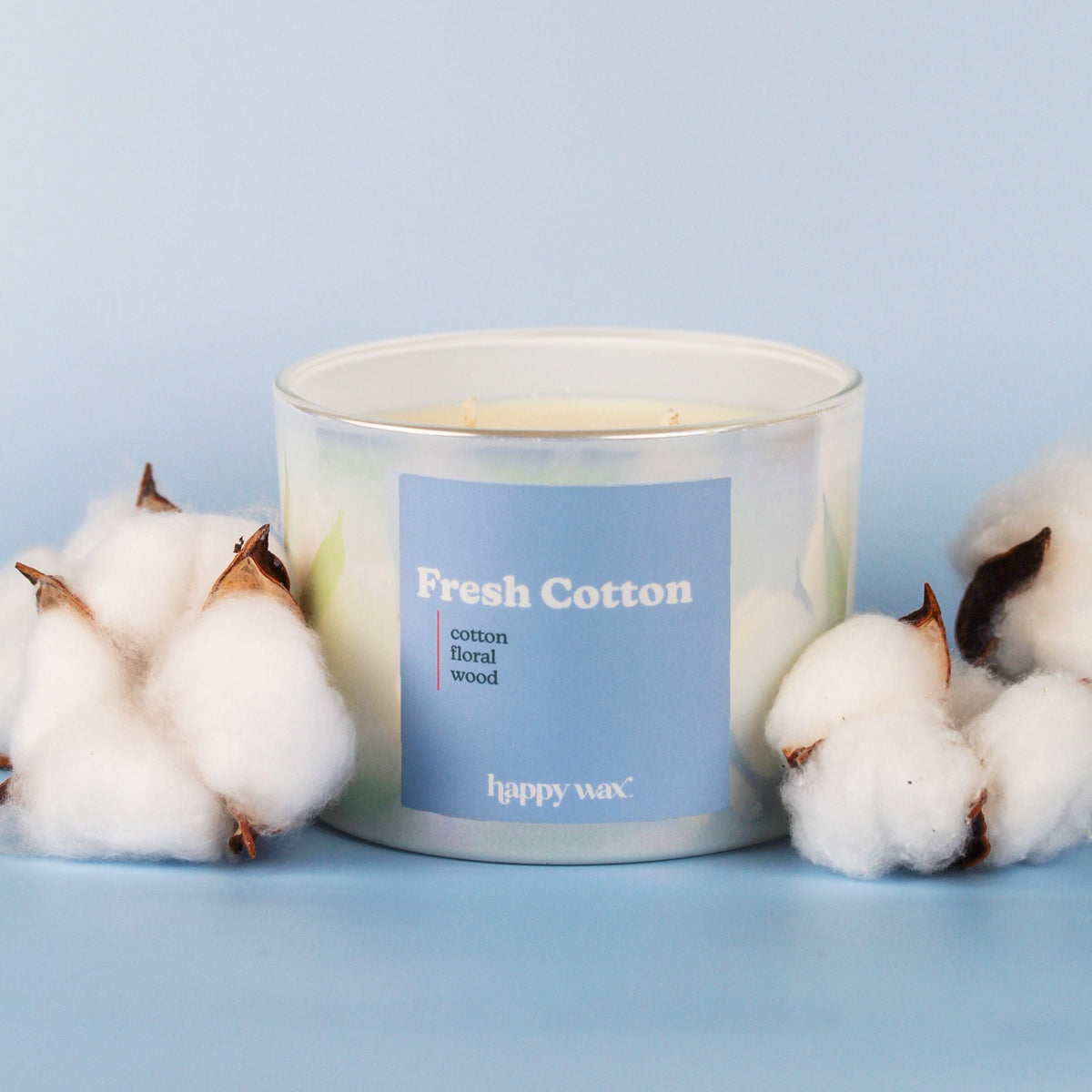 White three-wick candle with blue label surrounded by cotton flowers on light blue background.