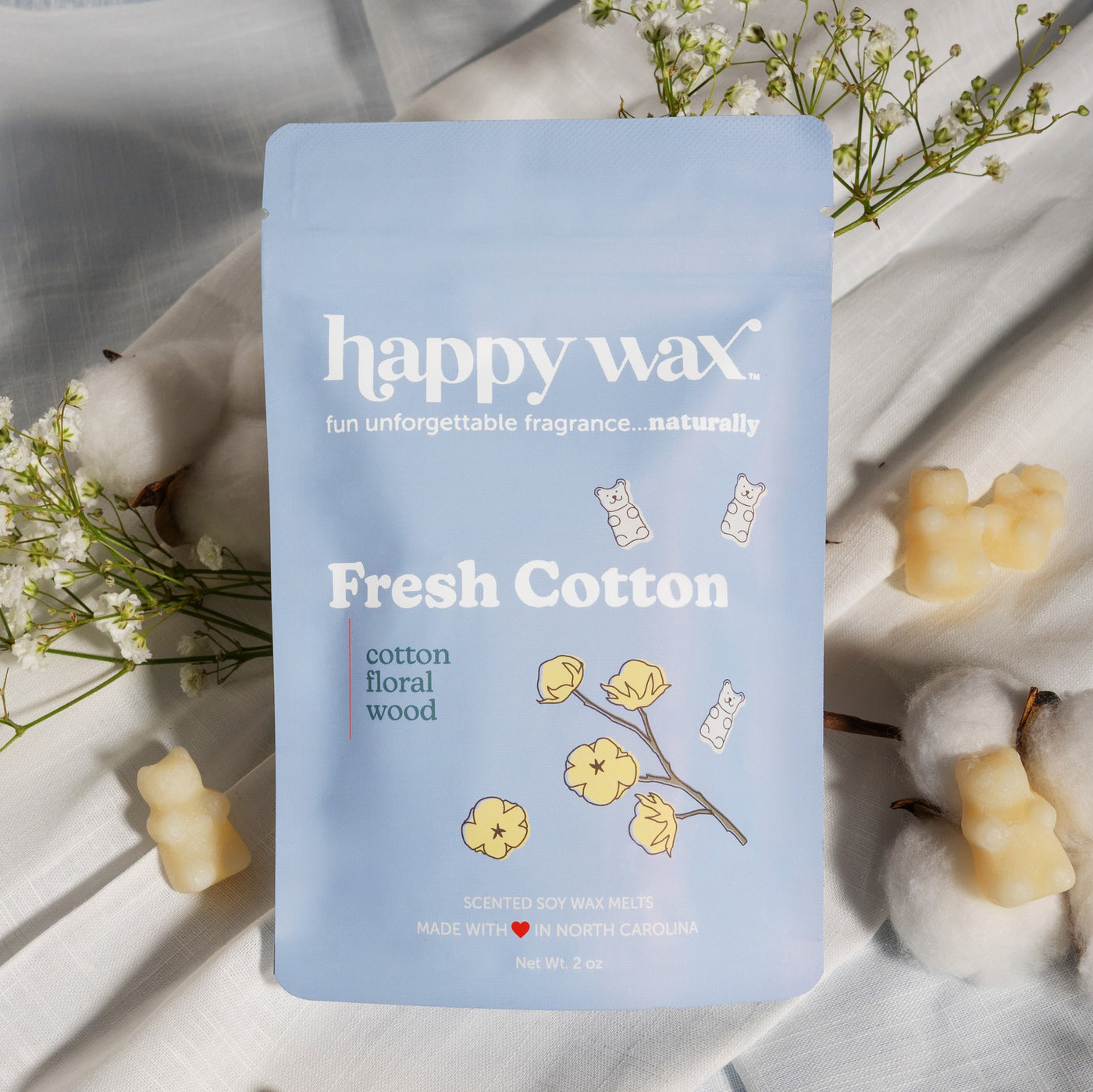 Light blue pouch of Fresh Cotton wax melts surrounded by pale yellow wax melts and baby's breath flowers on white fabric.