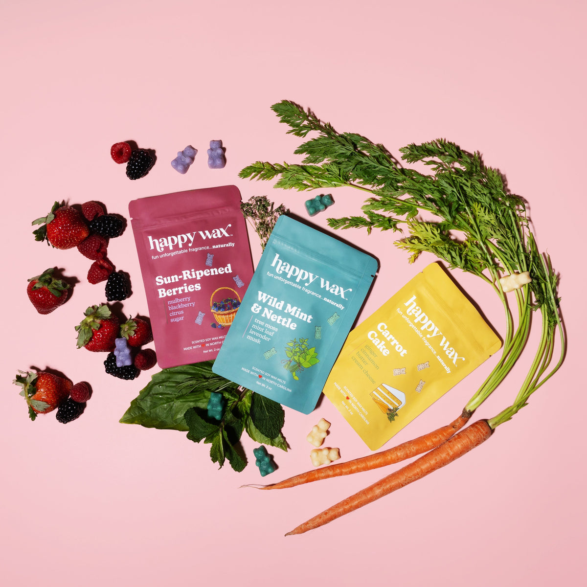 Three Happy Wax pouches in red, blue, and yellow with berries, herbs, and carrot on pink background.