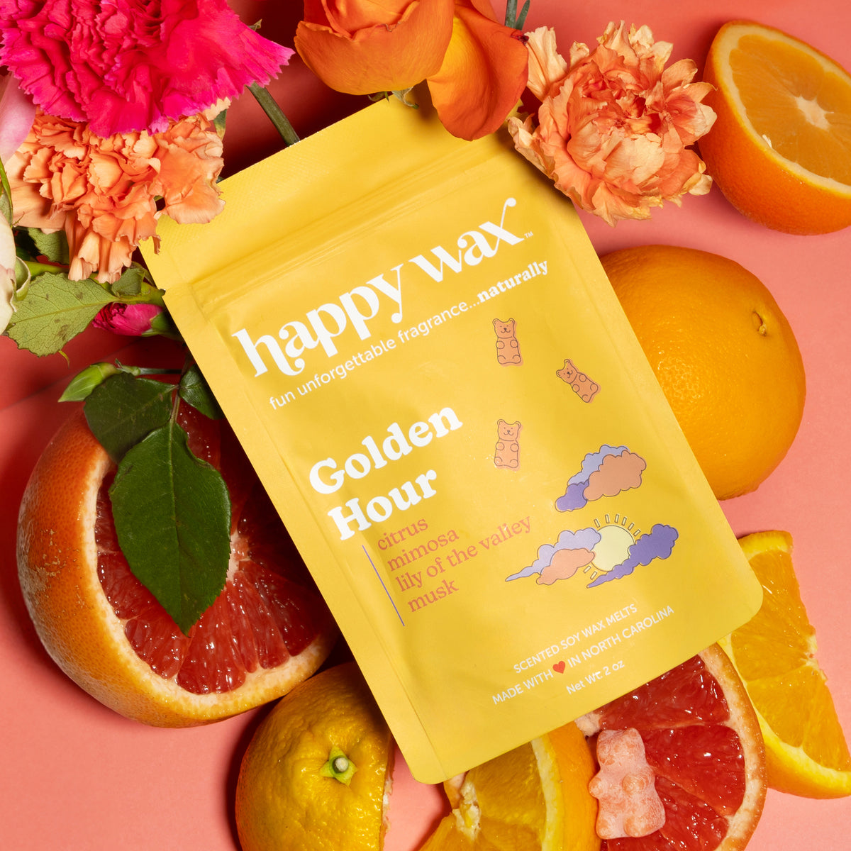Yellow wax melt pouch surrounded by citrus slices, grapefruit halves, orange flowers, and pink carnations on coral background