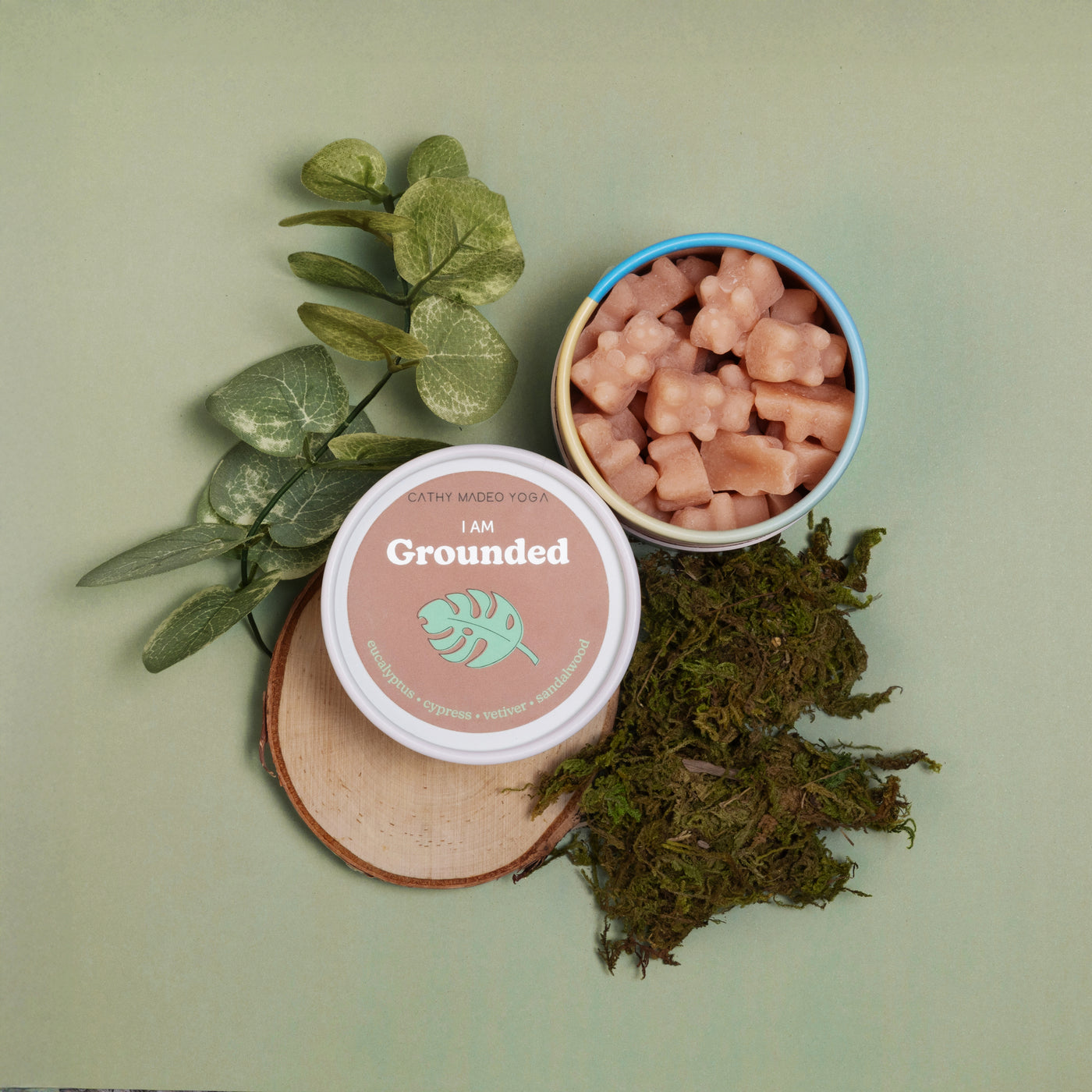 Pink wax melts in blue bowl with "I am Grounded" container, dried moss, and eucalyptus leaves on pale green background.