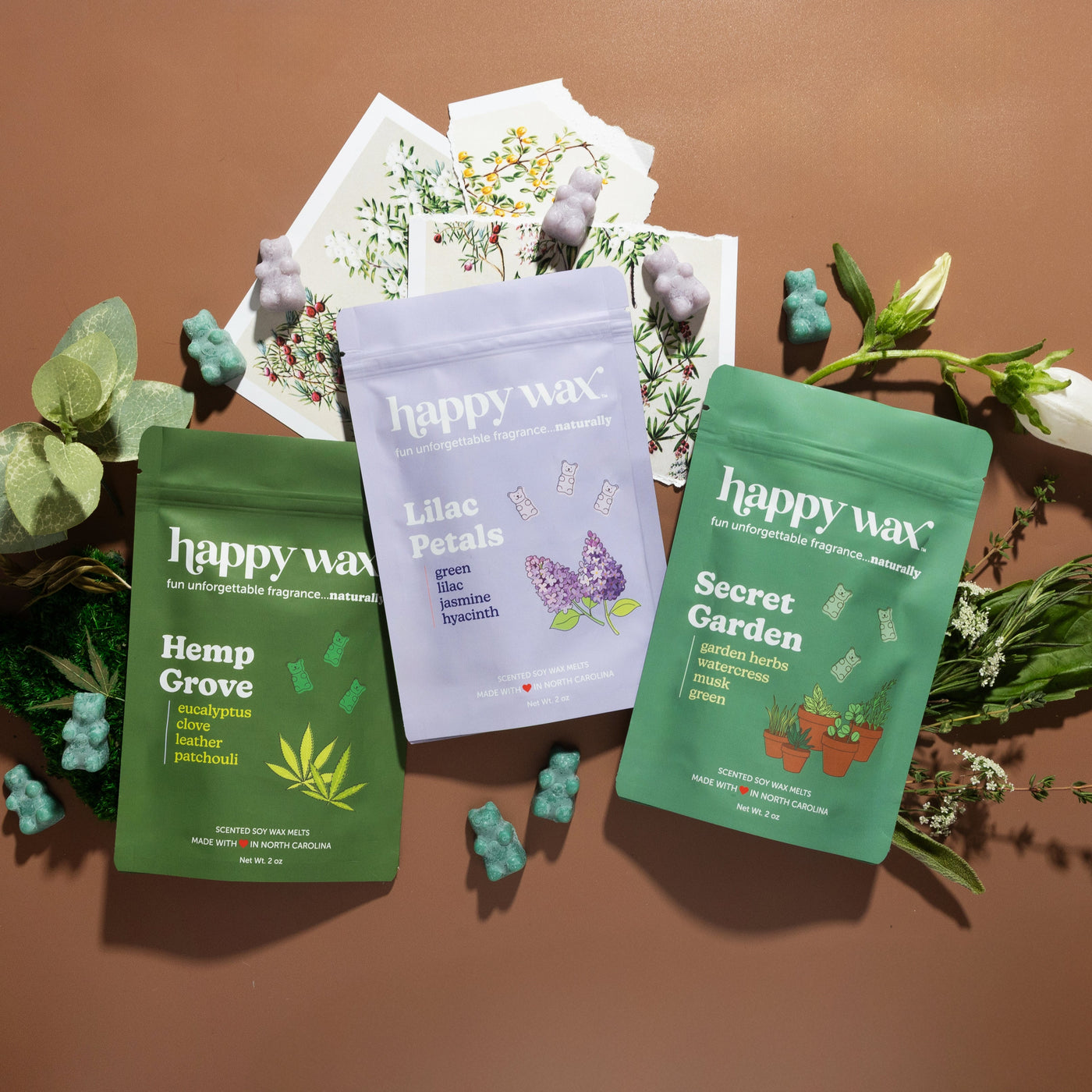 Three Happy Wax scented soy wax melt pouches in Hemp Grove, Lilac Petals, and Secret Garden flavors surrounded by botanical e