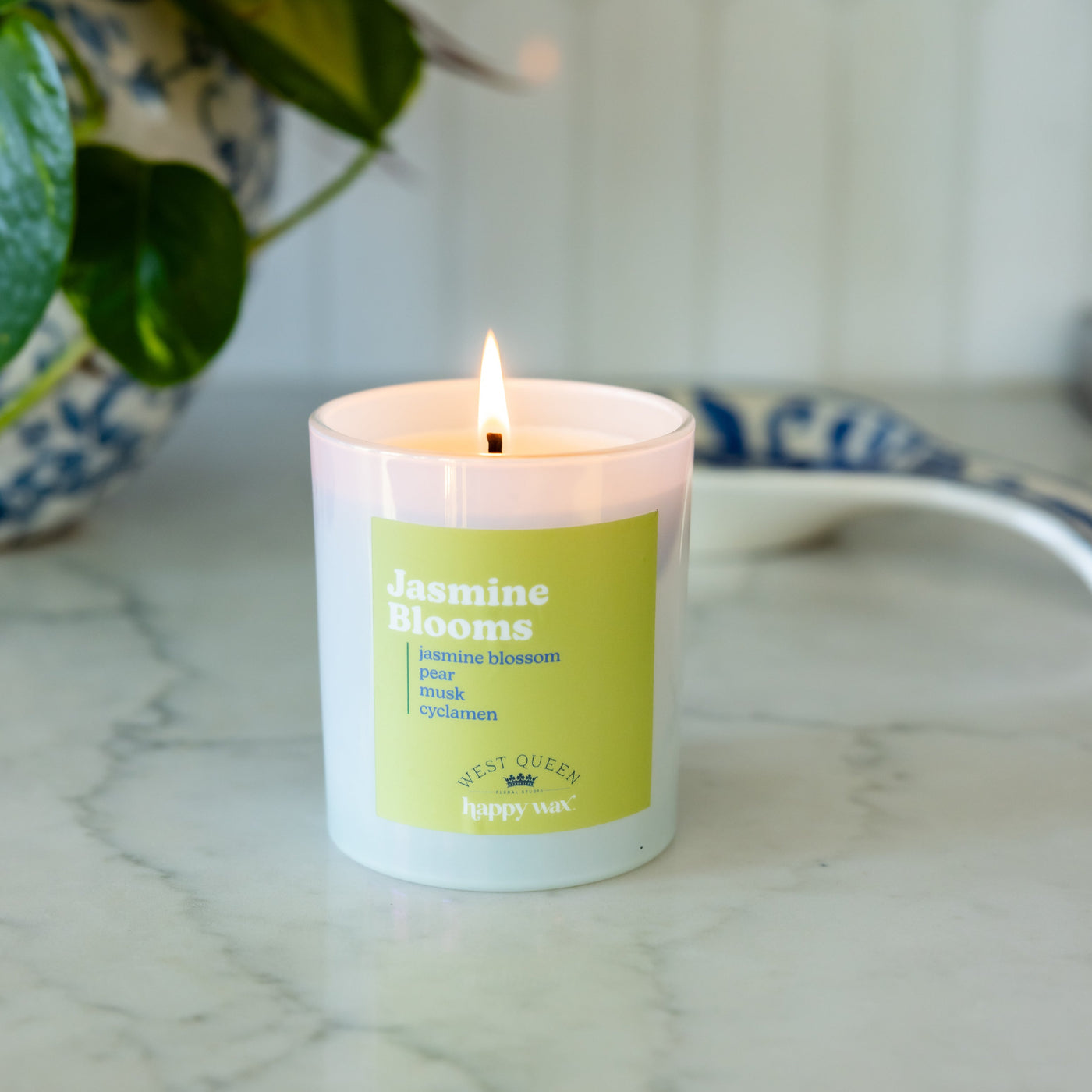 White jar candle with lime green label reading "Jasmine Blooms" and single lit wick, displayed on marble surface.