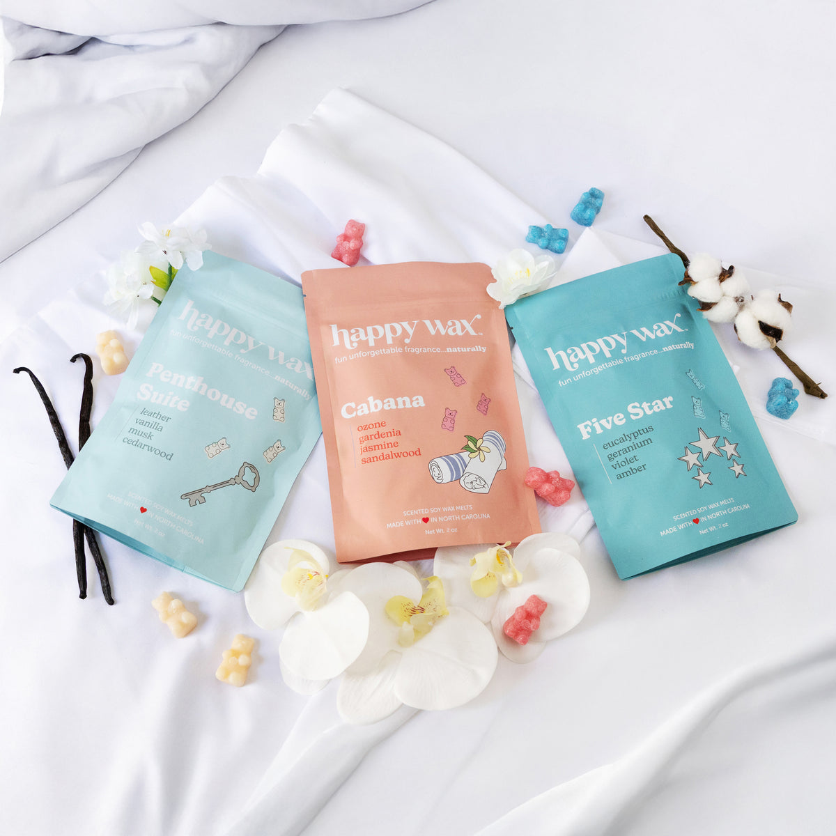 Three Happy Wax fragrance pouches in turquoise, coral, and turquoise with surrounding wax melts and natural décor elements.