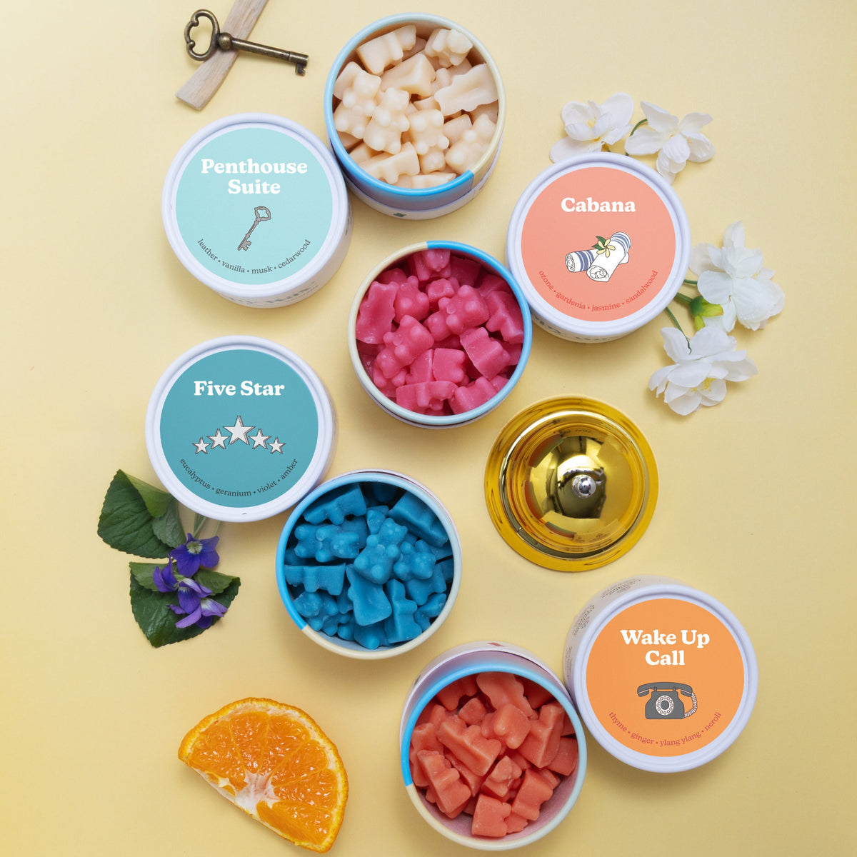 Four wax melts in labeled containers with scents like Penthouse Suite and Cabana, surrounded by decorative flowers and citrus