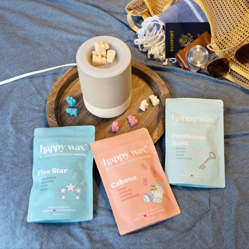 Three Happy Wax scented wax melt packets in blue, peach, and cream with a ceramic warmer containing wax cubes on a wooden tra