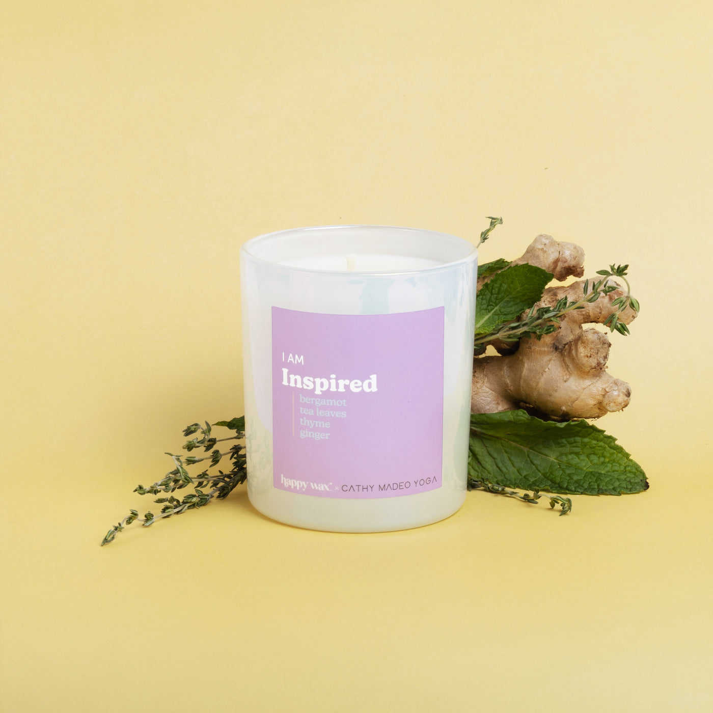 White cylindrical candle with pink label reading "I am Inspired" surrounded by ginger root and green leaves on yellow backgro
