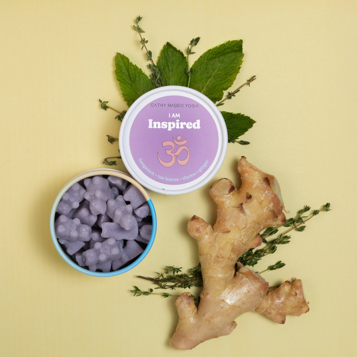 Purple wax melts in blue container with "I am Inspired" lid, surrounded by fresh mint leaves and ginger root on pale yellow b