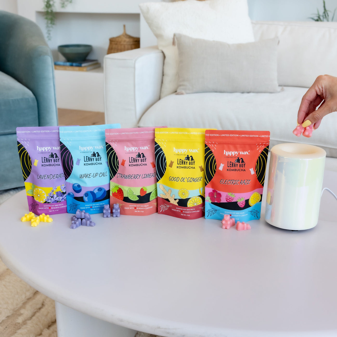 Five colorful kombucha pouches in lavender, blueberry, strawberry, ginger, and grapefruit flavors with matching wax melts dis