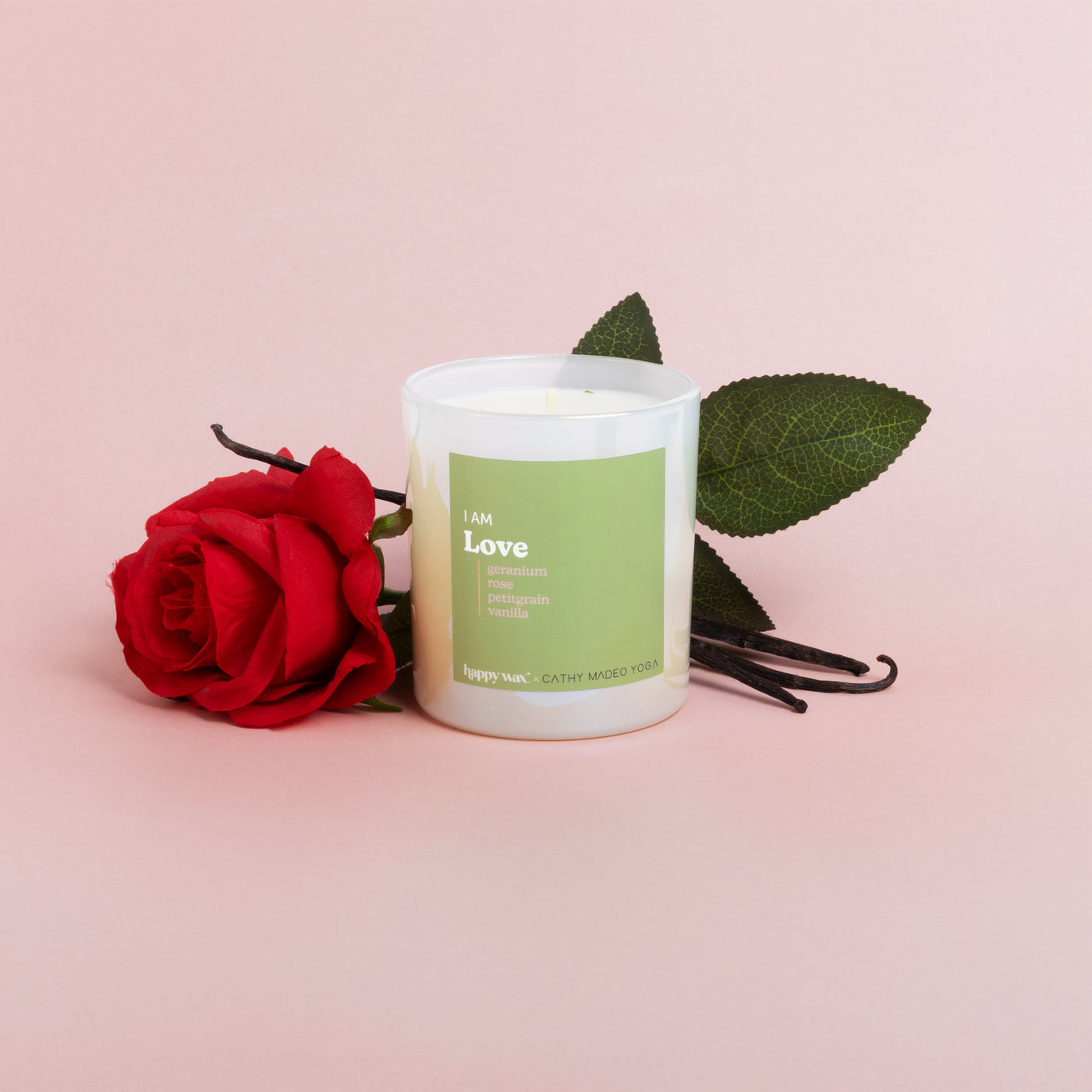 White cylindrical candle with green label surrounded by red rose, vanilla pods, and green leaves on pink background.