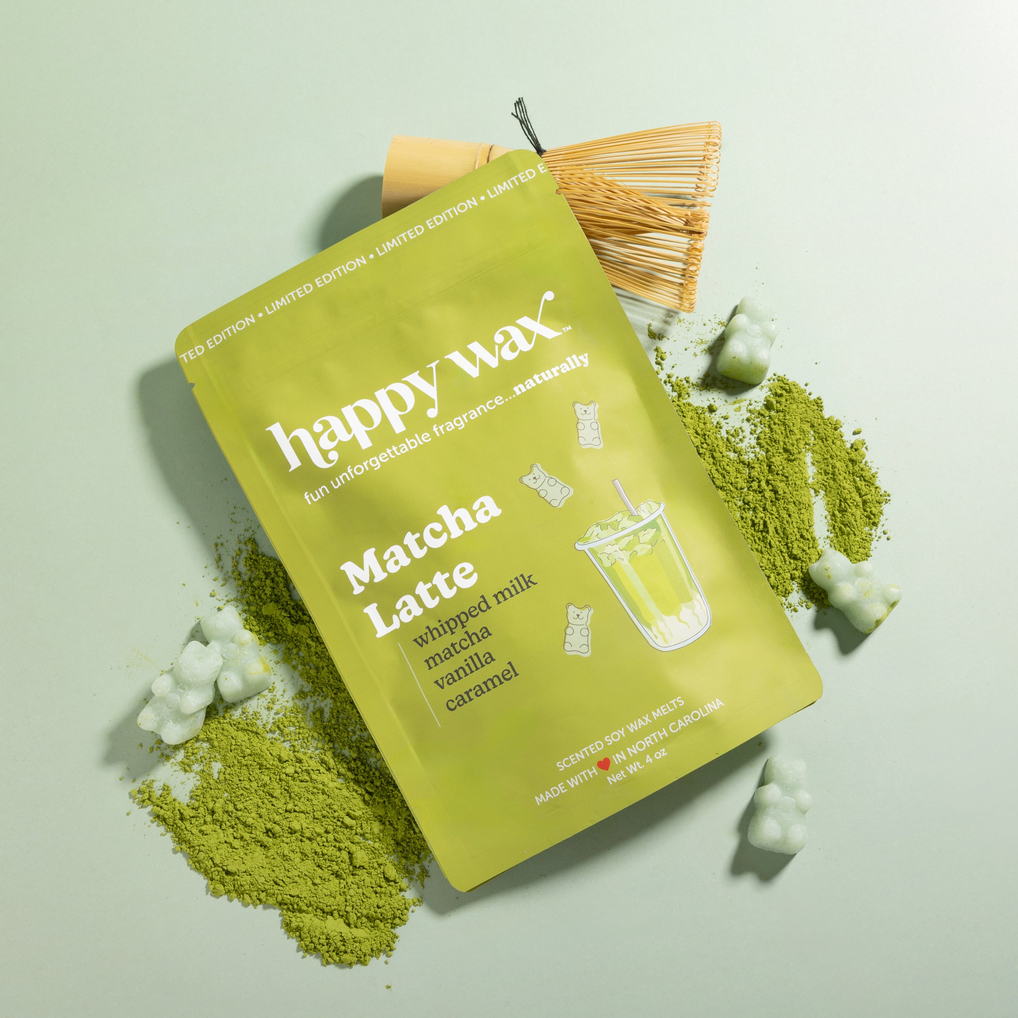 Lime green wax box labeled "Matcha Latte" surrounded by matcha powder, a whisk, and decorative wax pieces on pale background.