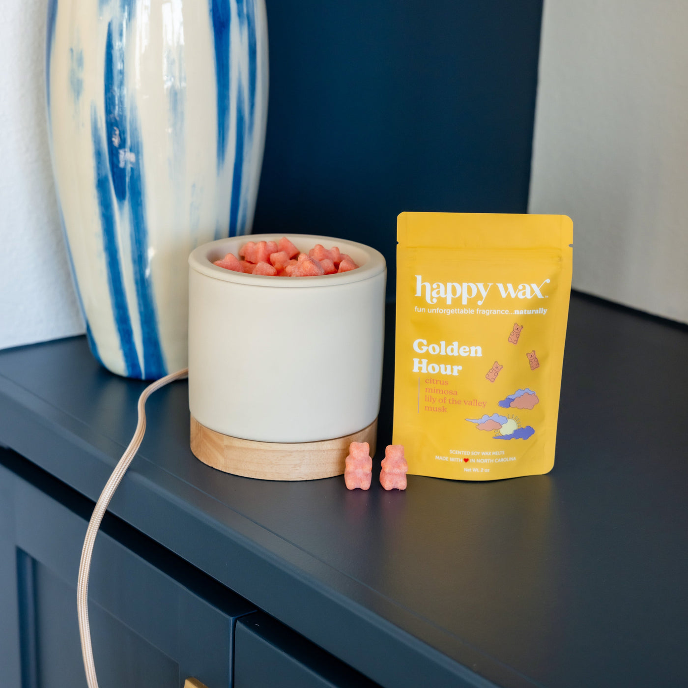 White wax warmer with wooden base, pink wax melts inside, yellow Happy Wax pouch, and two pink wax bears displayed on dark su