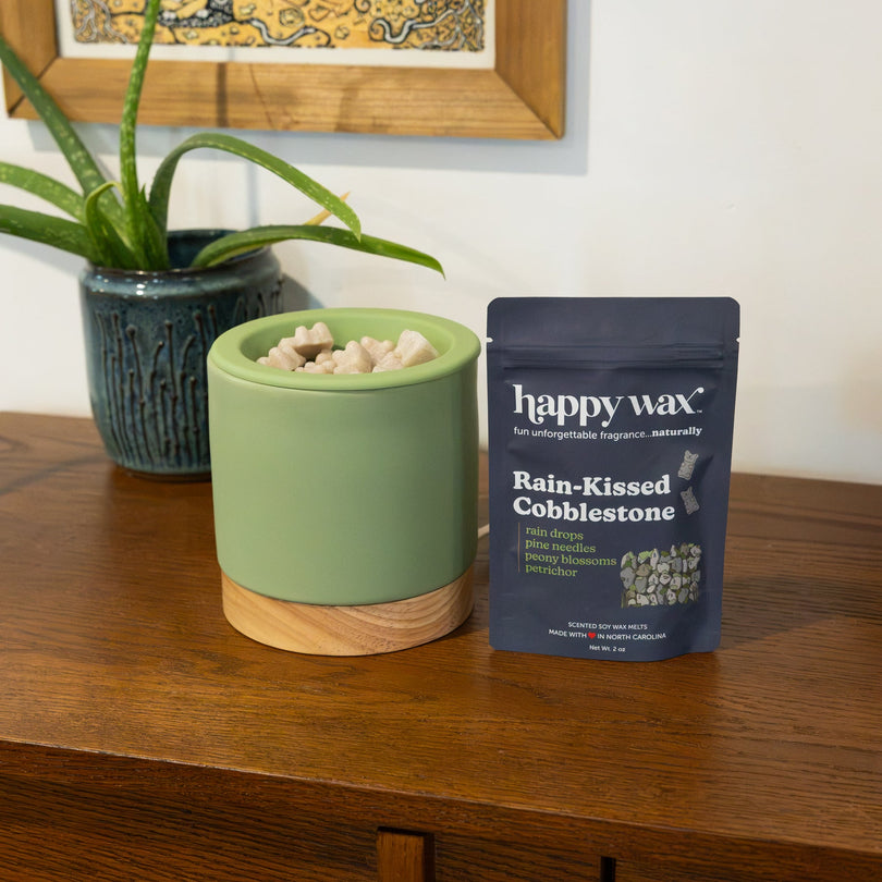Green ceramic wax warmer with wooden base filled with wax melts, displayed next to Happy Wax fragrance pouch on wooden table.