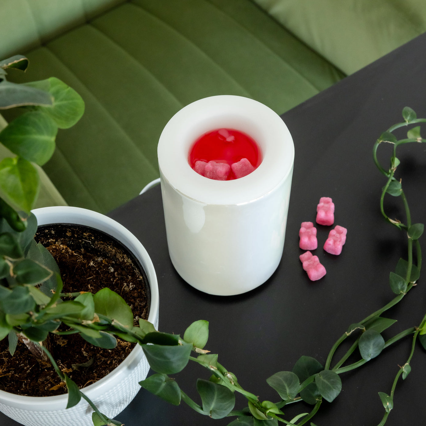 White cylindrical wax warmer with red wax and pink wax cubes on dark surface surrounded by potted plants.