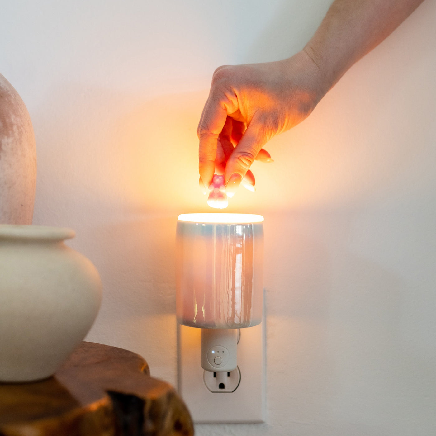 Hand holding iridescent wax warmer with glowing orange light, mounted on wall outlet with white base.