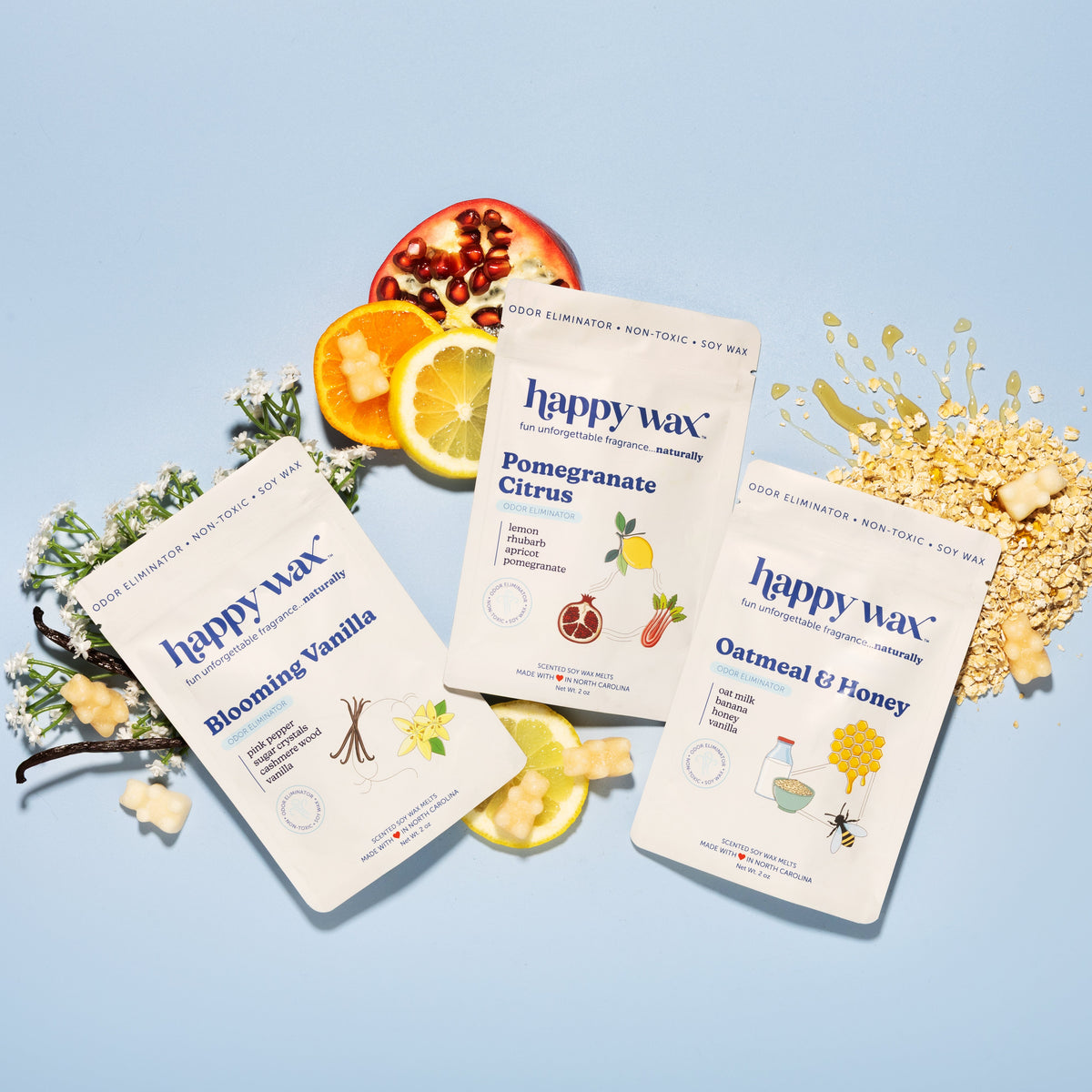 Three cream-colored soy wax candle packages labeled Blooming Vanilla, Pomegranate Citrus, and Oatmeal & Honey, arranged with