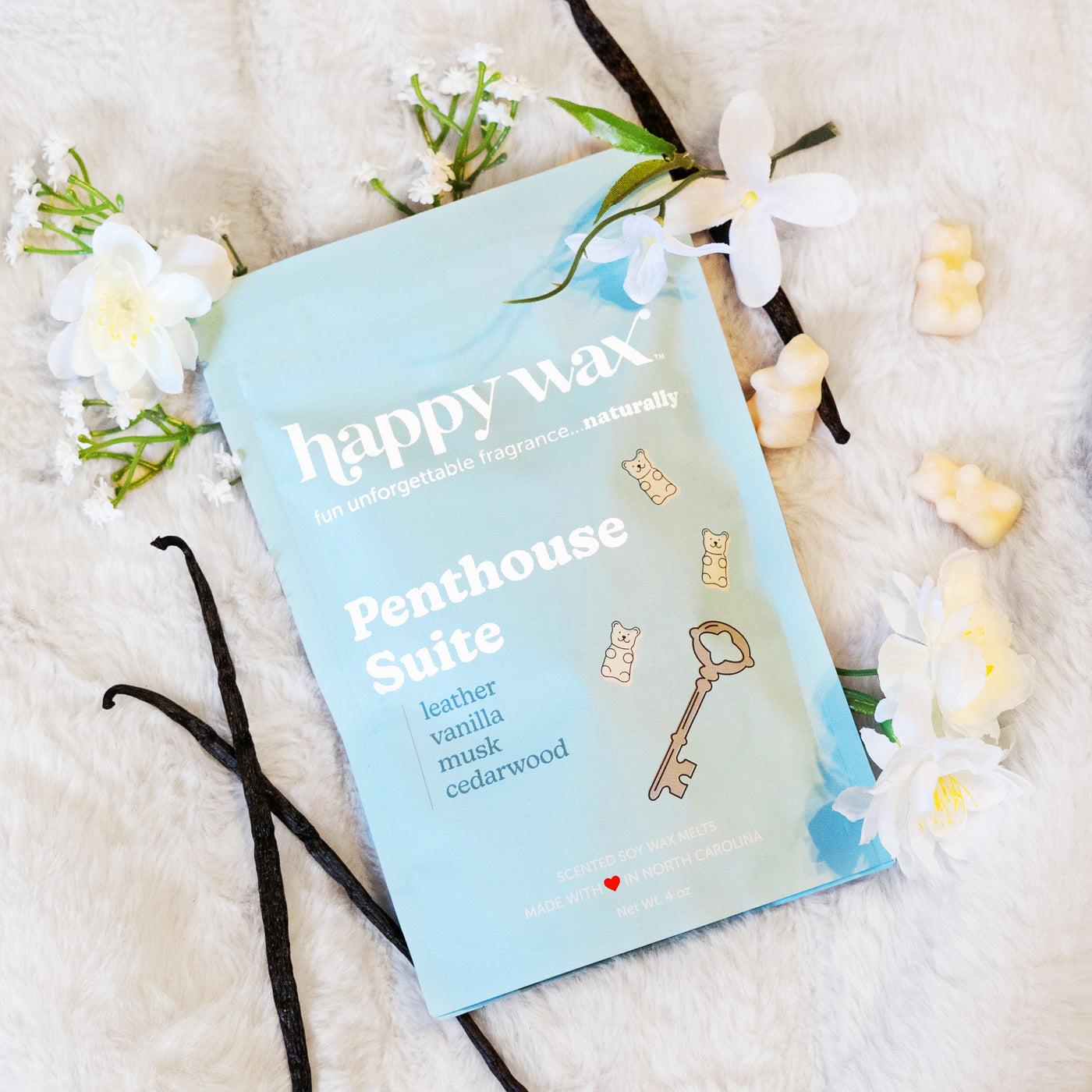 Light blue wax melt package labeled "Penthouse Suite" surrounded by cream-colored wax melts, white flowers, and vanilla beans