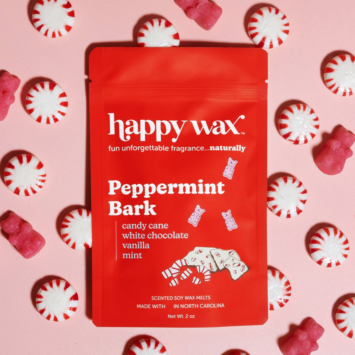 Red Happy Wax pouch of peppermint bark scented soy wax melts surrounded by candy canes and pink gummy bears on pink backgroun