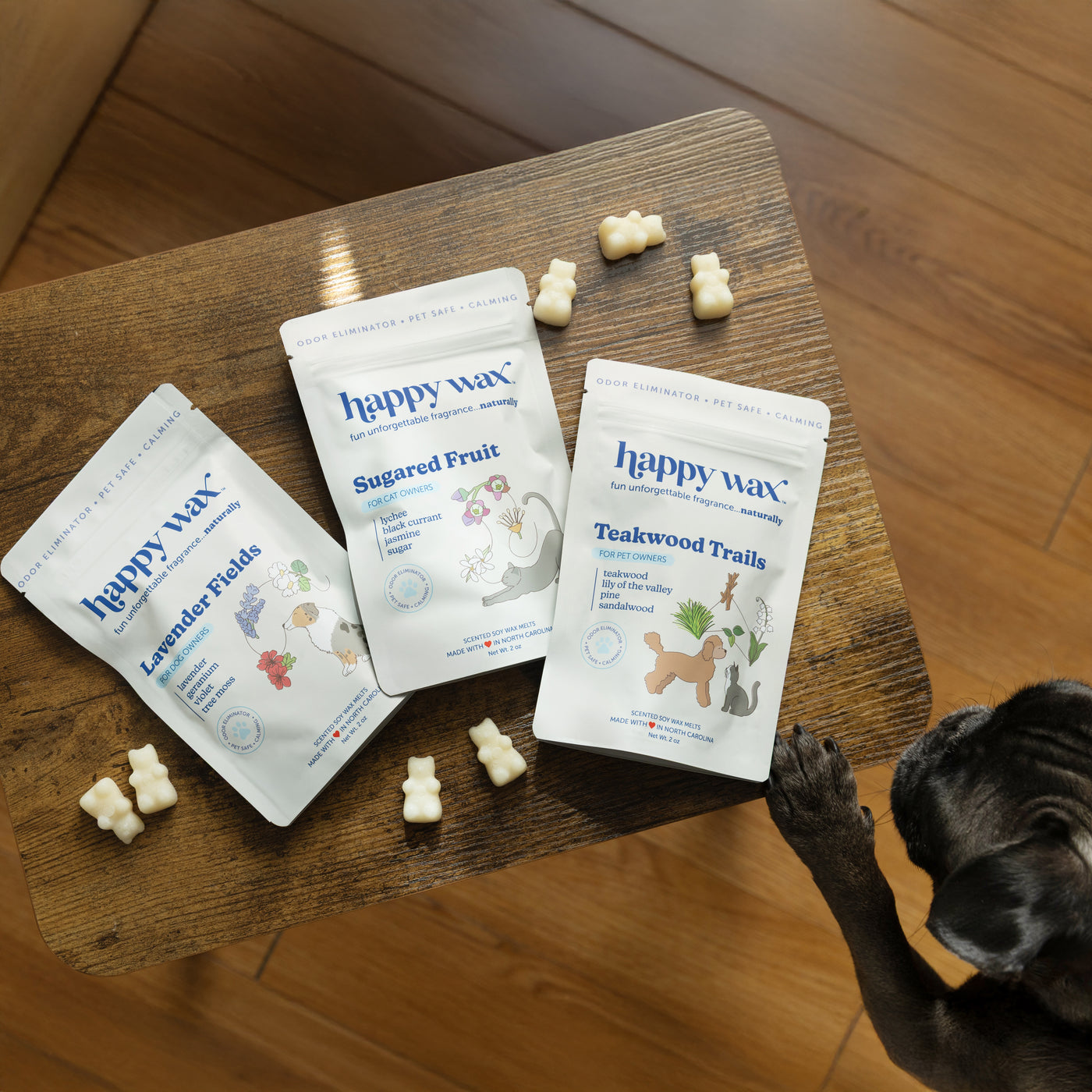 Three Happy Wax pet product pouches in Lavender Fields, Sugared Fruit, and Teakwood Trails scents with wax melts on wooden su