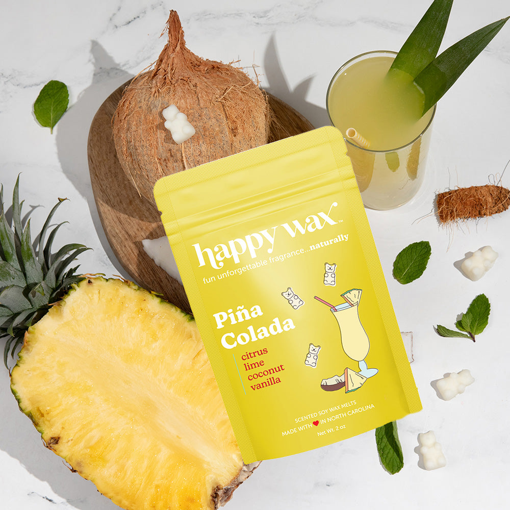 Yellow Pina Colada wax melt pouch surrounded by pineapple, coconut, tropical drink, and mint leaves on marble background.