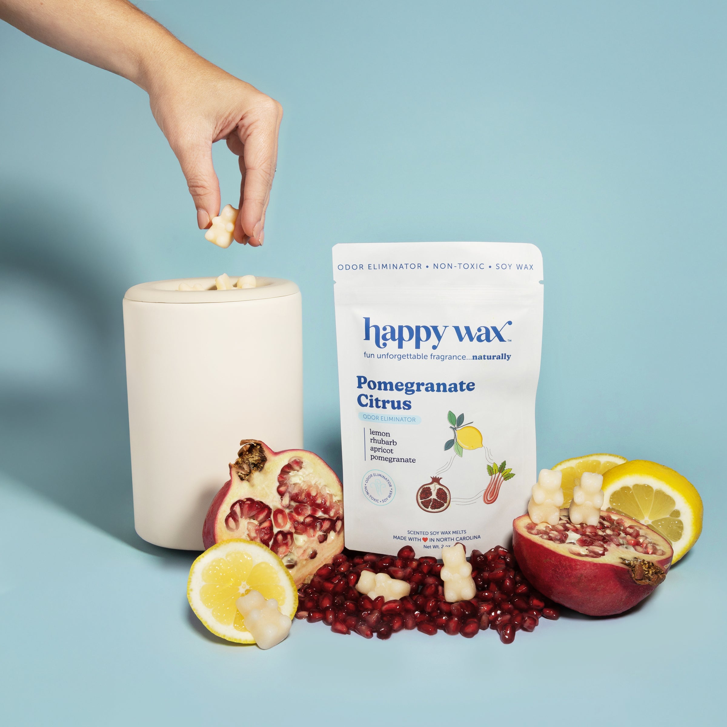 Hand holding wax melt over white ceramic warmer with Happy Wax pomegranate citrus package and fresh fruit.