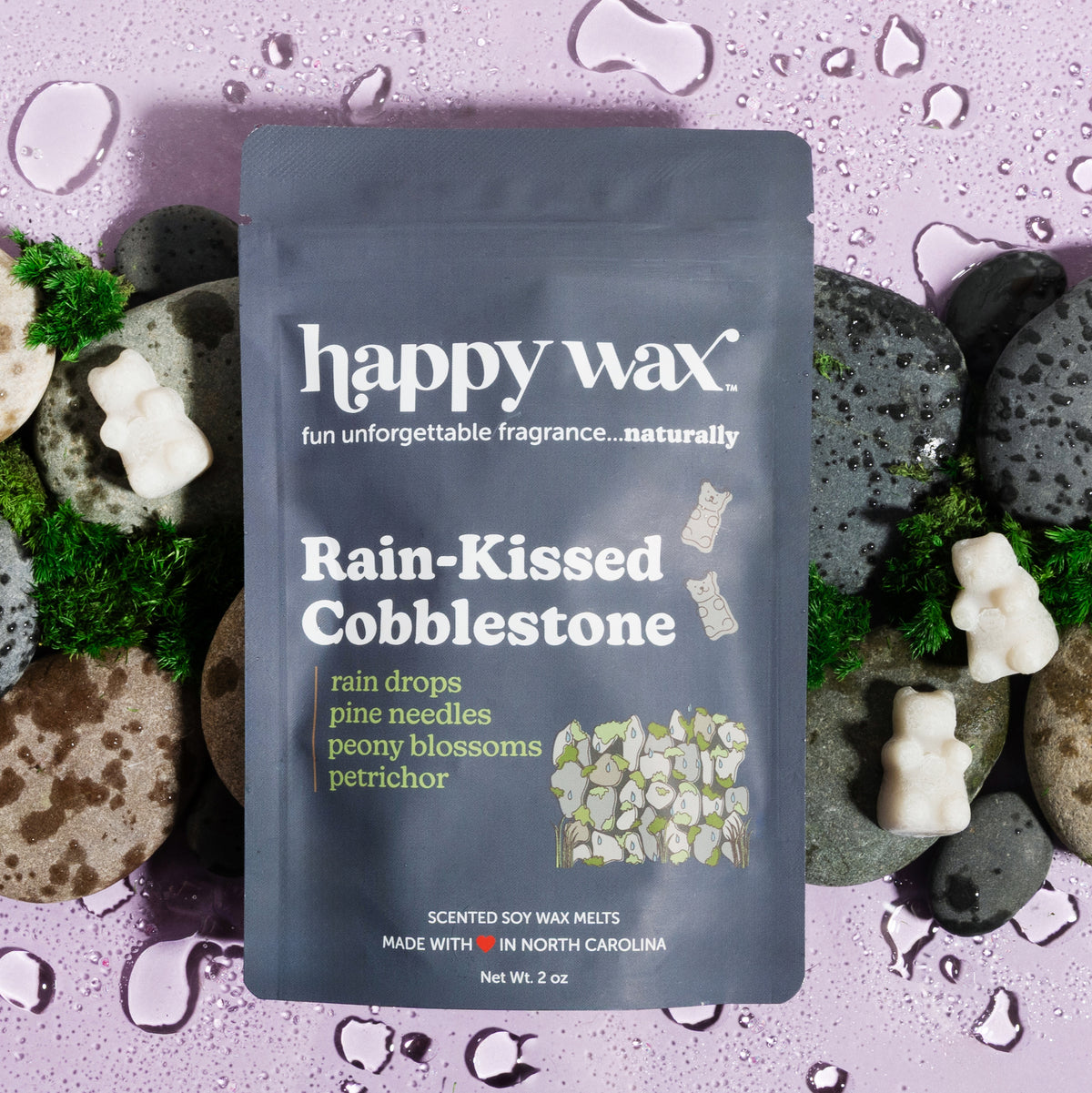 Dark blue pouch of happy wax soy wax melts surrounded by white wax melts, moss, and porous stones on purple speckled backgrou