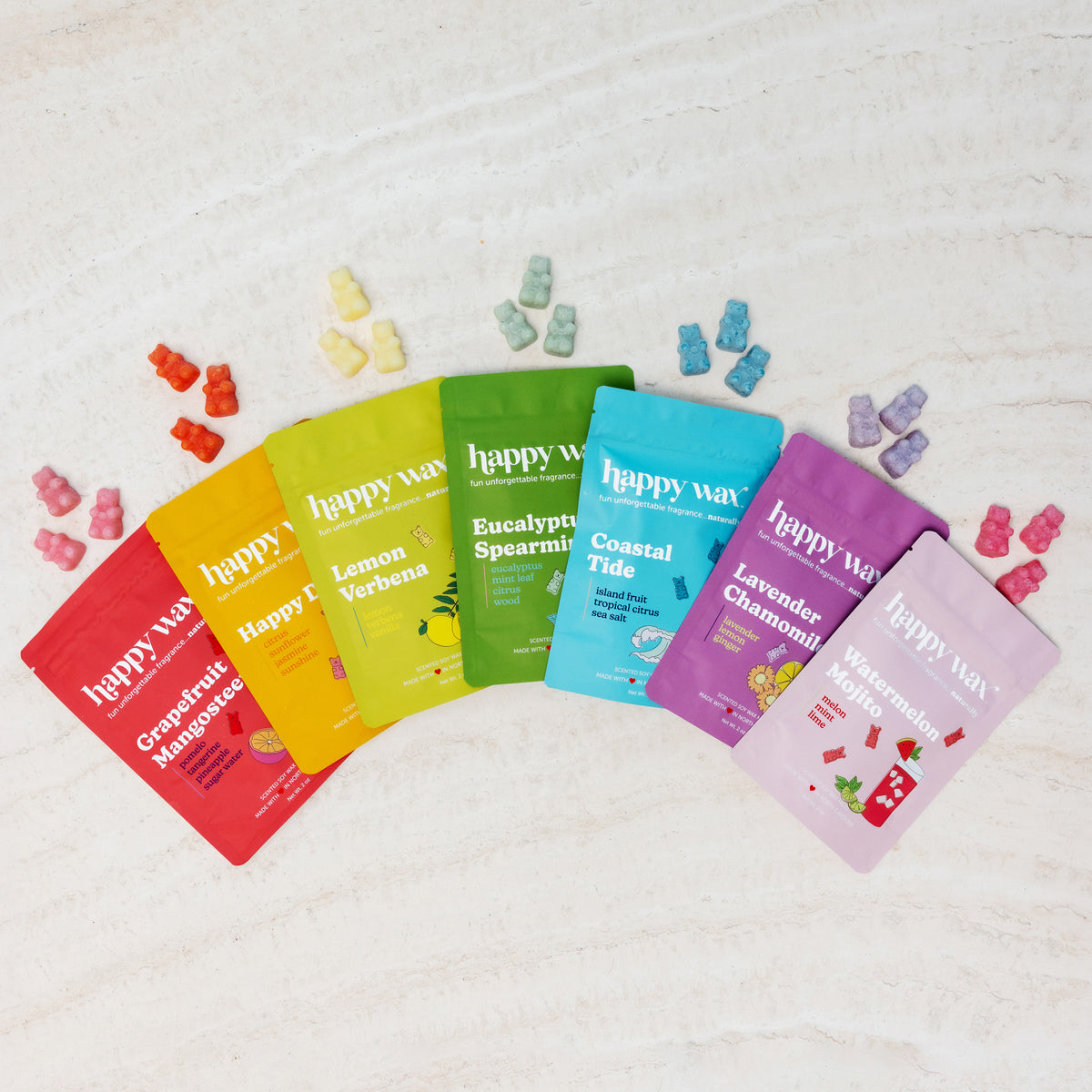 Seven colorful wax melts packages arranged in a rainbow arc with coordinating scented wax cubes displayed above on white wood