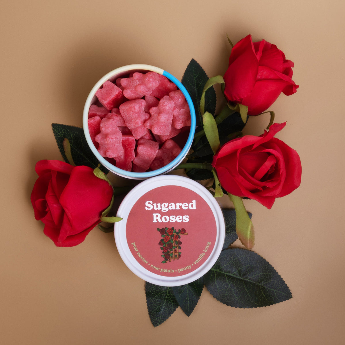 Pink wax melts in white container with blue rim surrounded by fresh red roses and green leaves on tan background.