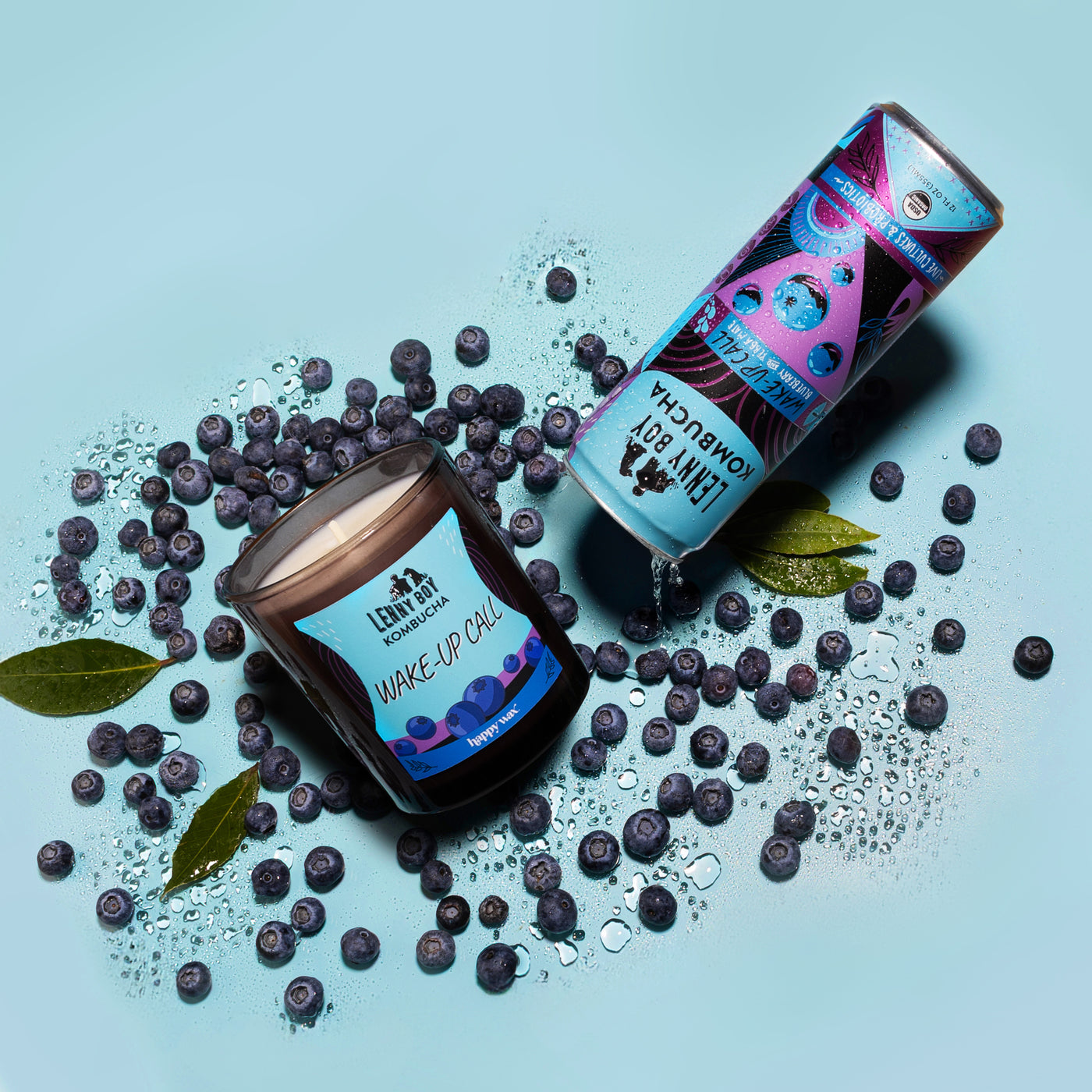 Jar and tube candle surrounded by blueberries and green leaves on light blue background.