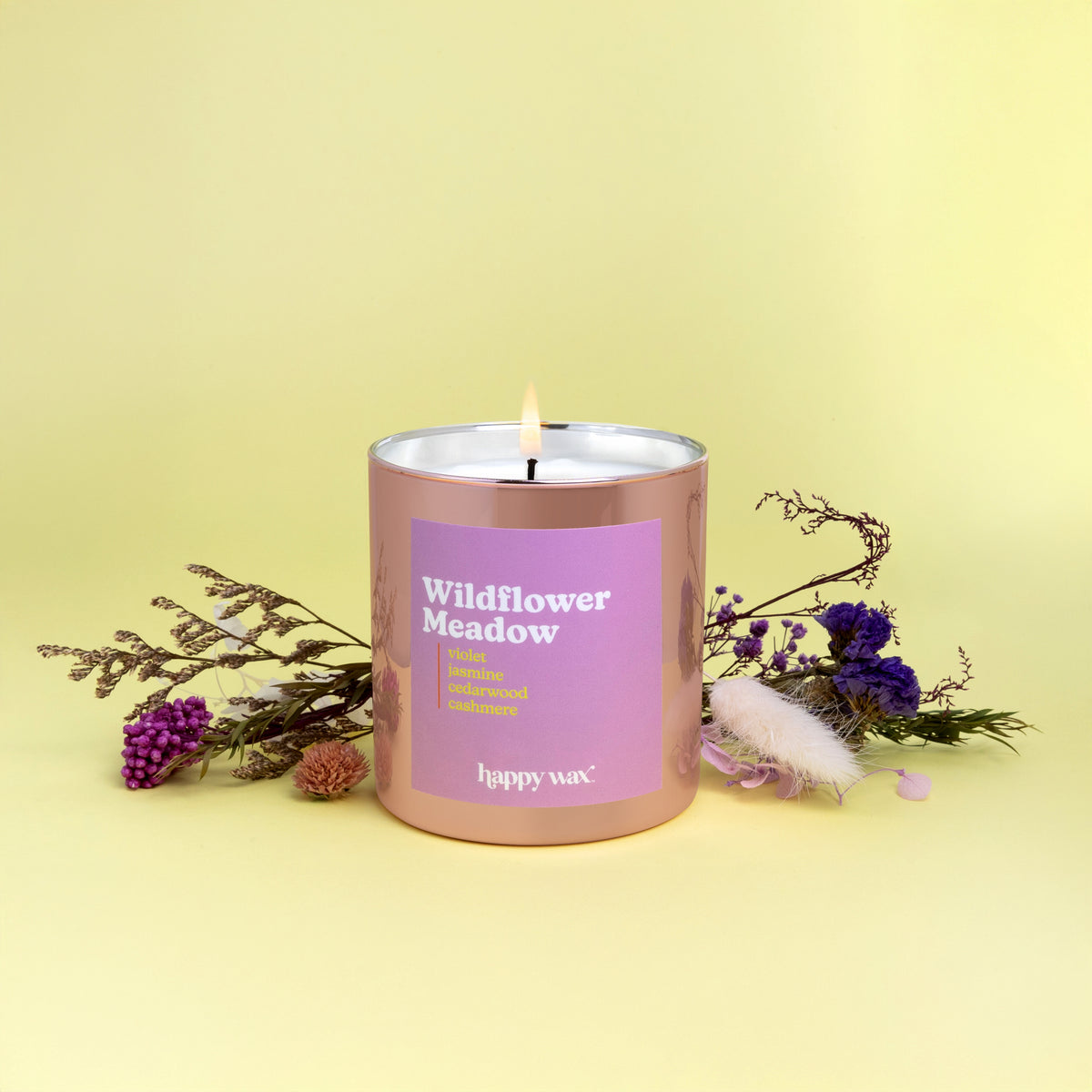 Rose gold cylindrical candle with purple label and single lit wick, surrounded by dried wildflowers on yellow background.