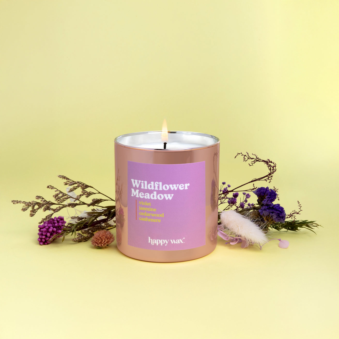 Rose gold cylindrical candle with purple label and single lit wick, surrounded by dried wildflowers on yellow background.