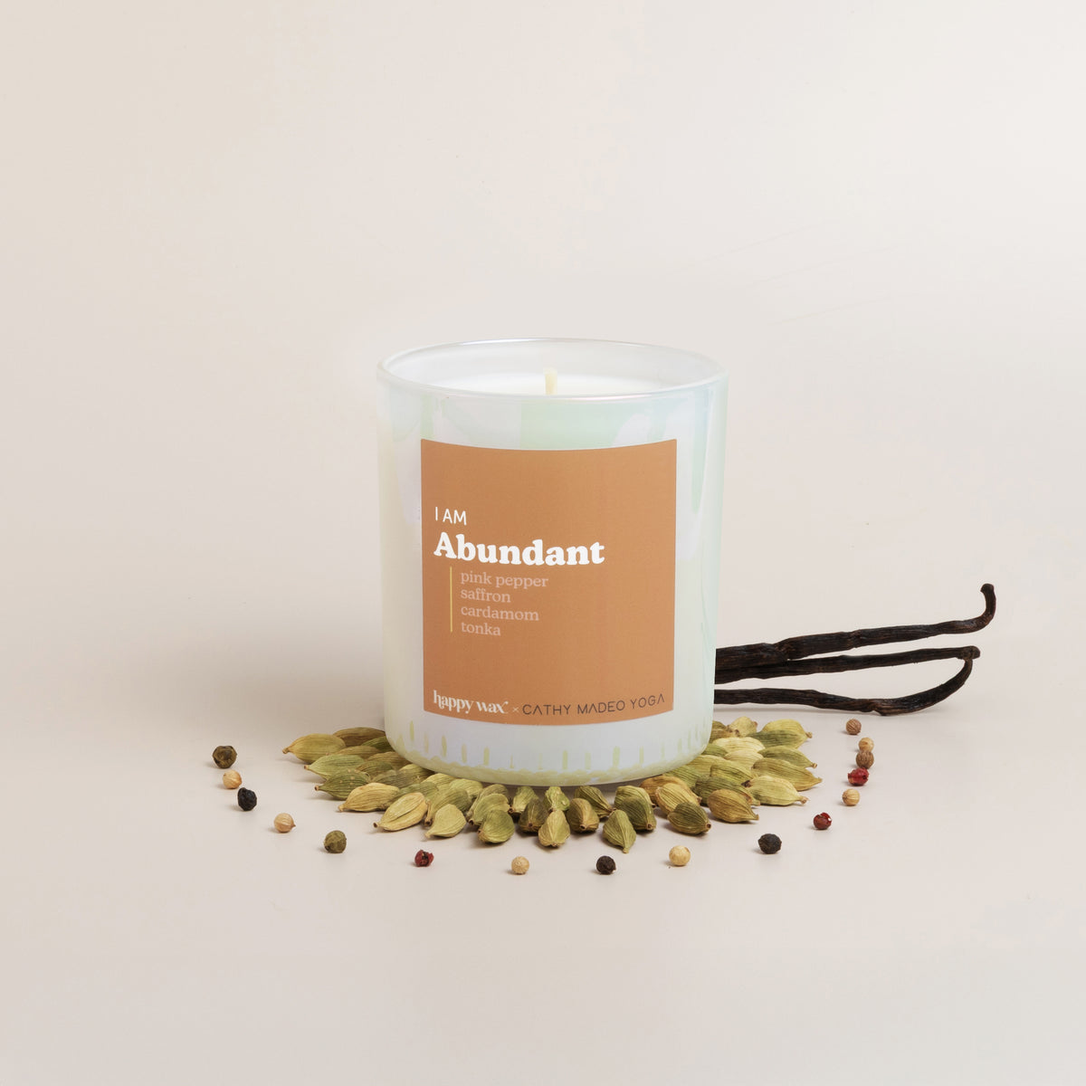Clear glass candle with tan label reading "I am Abundant," surrounded by cardamom pods and peppercorns, with vanilla bean pod