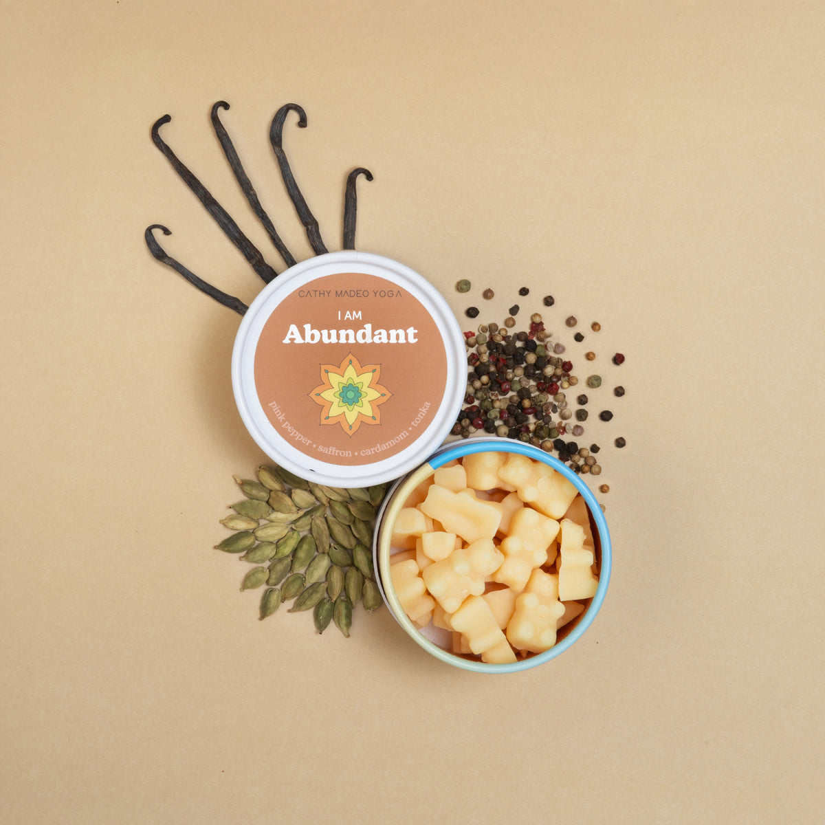 Two containers of "I am Abundant" wax melts with cream-colored wax pieces, vanilla beans, peppercorns, and dried herbs on bei