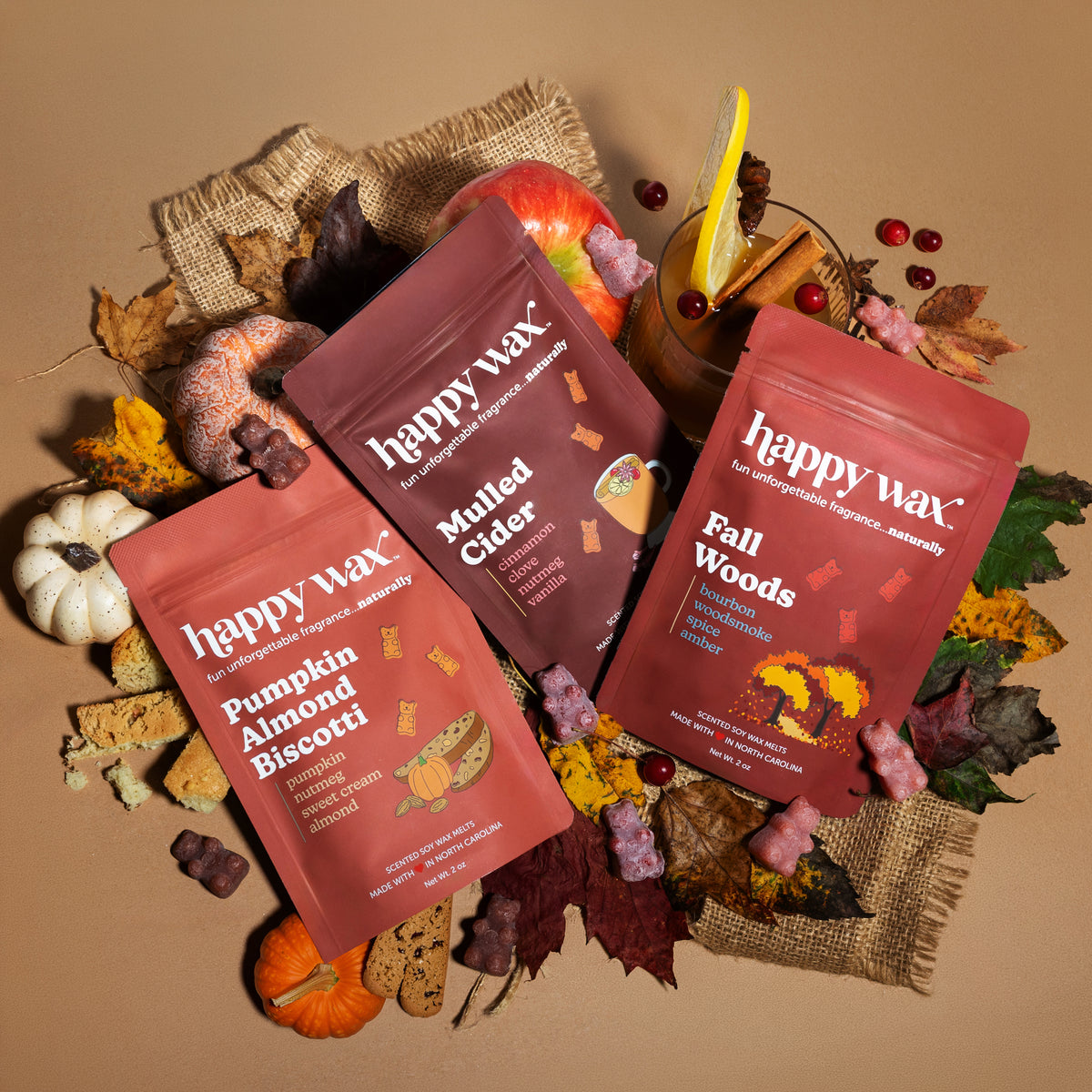 Three happy wax pouches in burgundy and coral with Fall Woods, Mulled Cider, and Pumpkin Birch scents surrounded by autumn leaves, cider, pumpkins and burlap