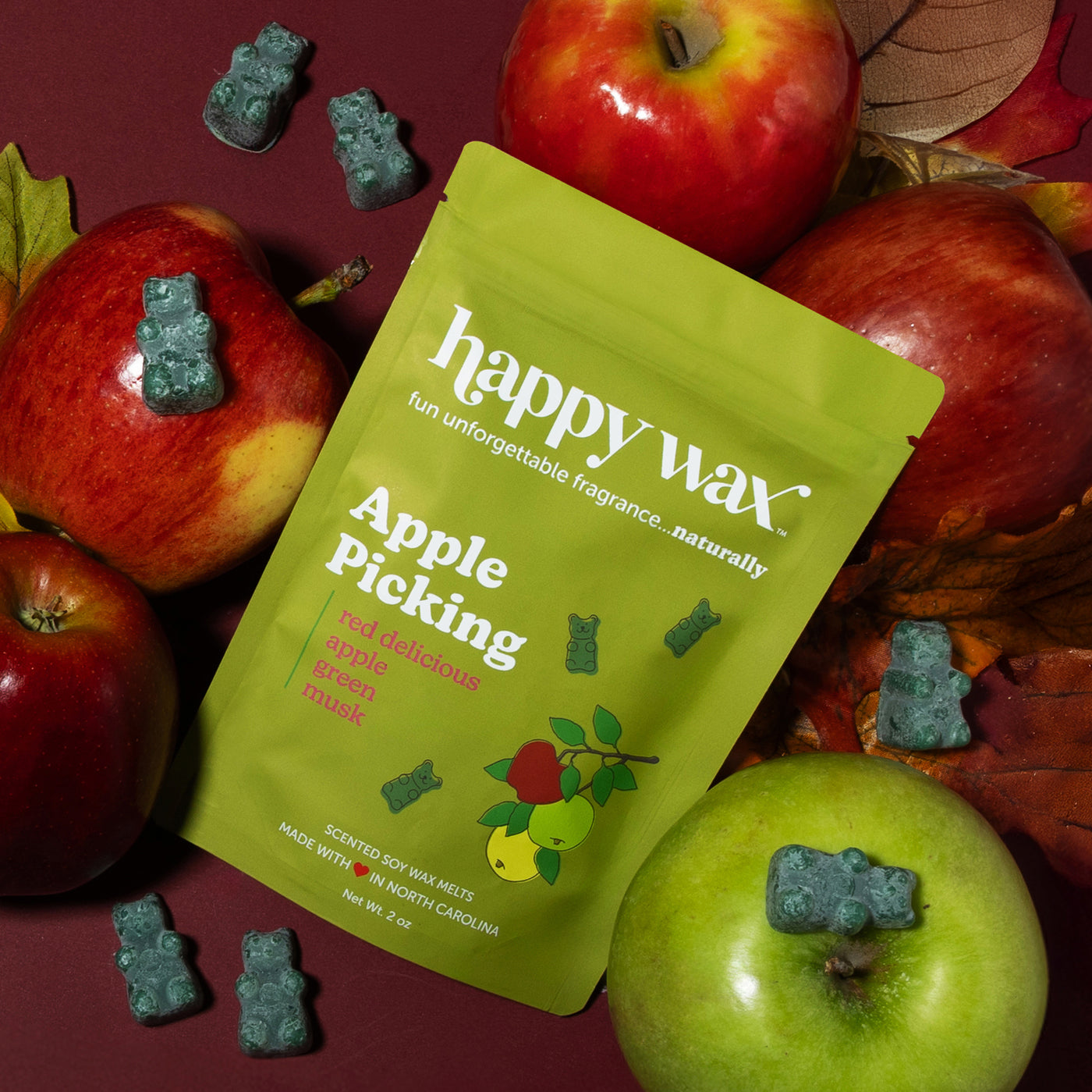 Lime green wax melt pouch surrounded by fresh red and green apples and teal bear-shaped wax melts on burgundy background.