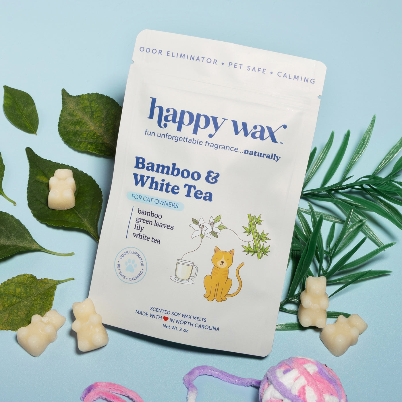 White package of bamboo and white tea scented wax melts surrounded by green leaves and cream-colored wax pieces on light blue